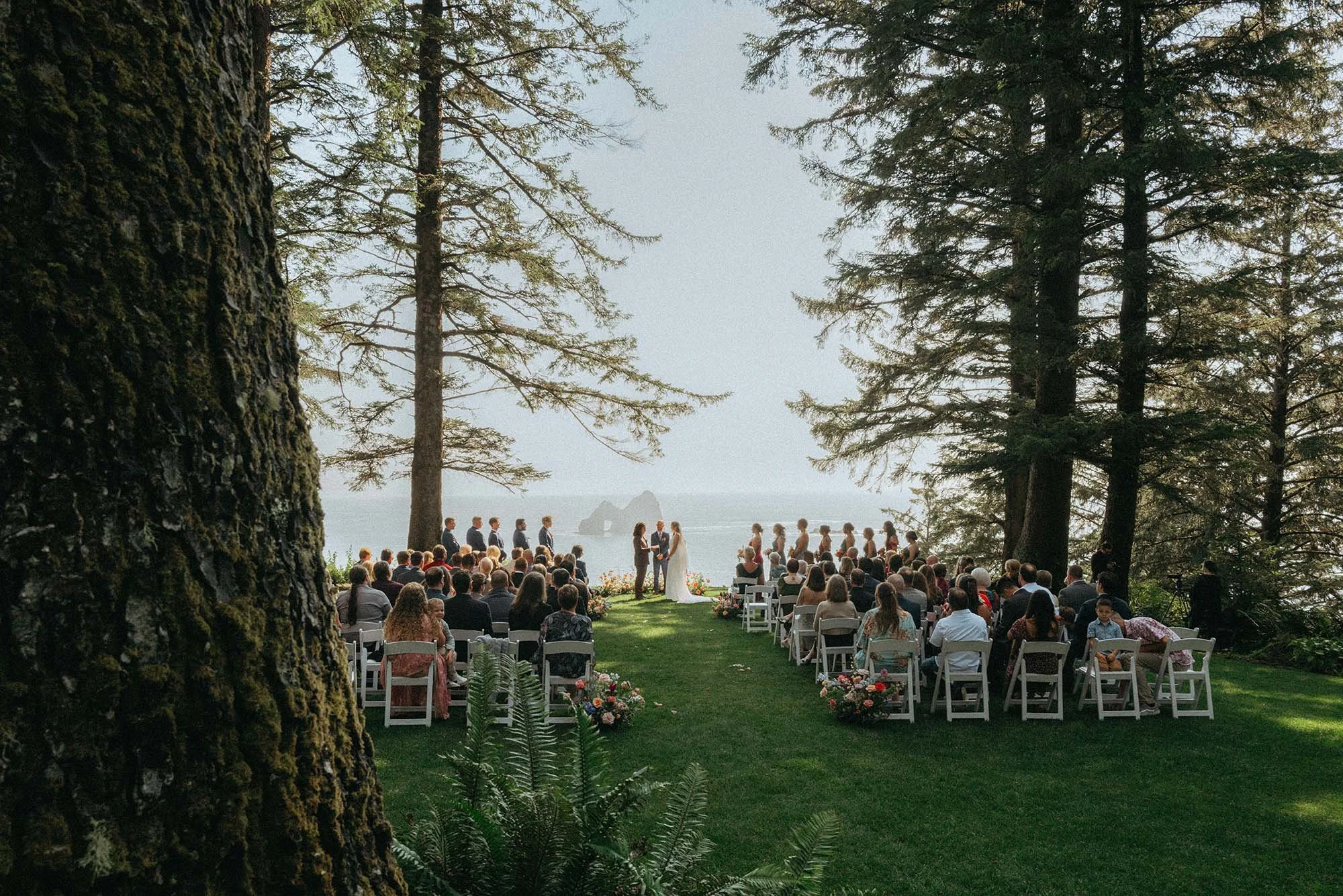 Outdoor wedding ceremony on grassy area by the water, with guests seated on white chairs, a bride and groom exchanging vows, and a backdrop of large trees and ocean.