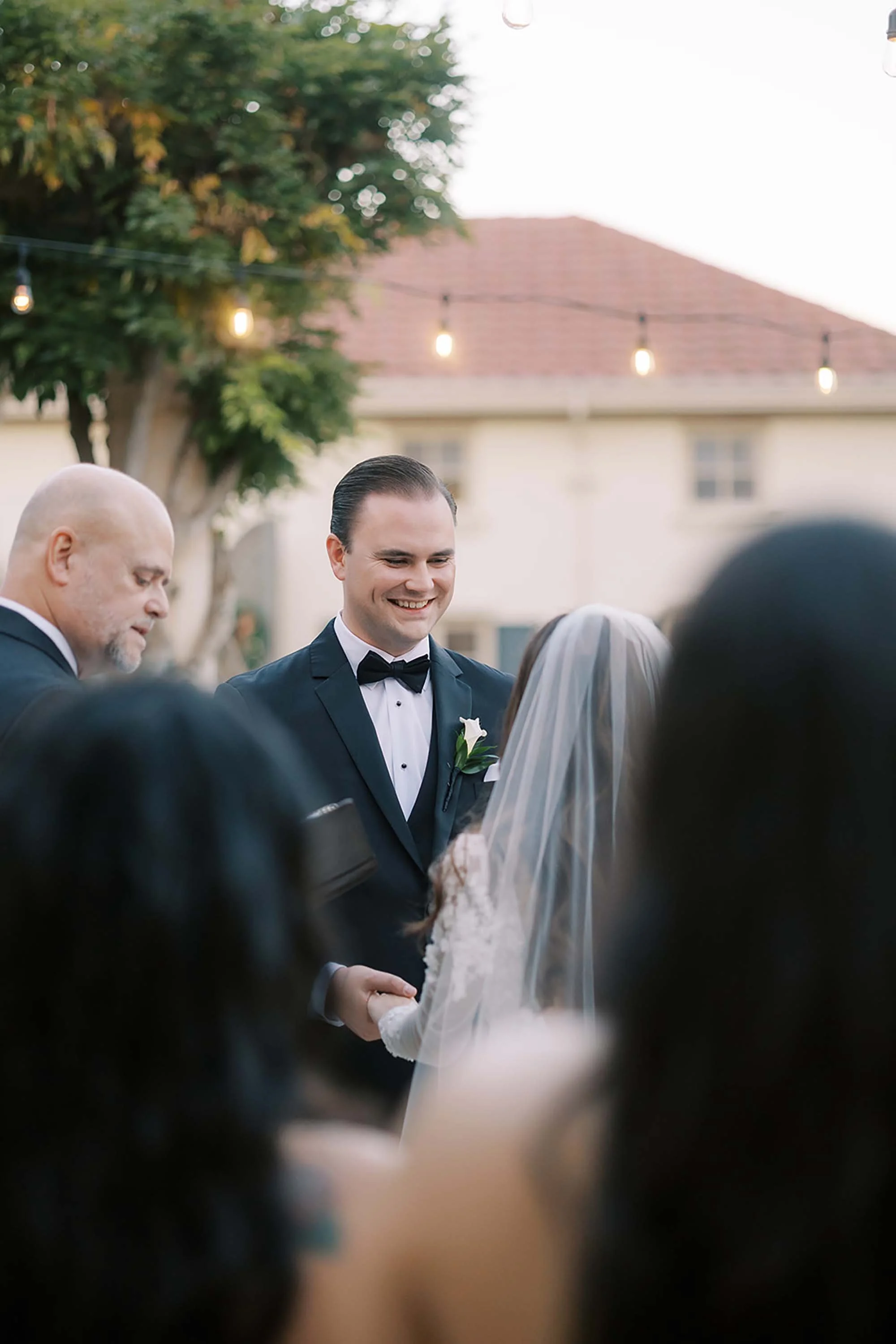 A wedding ceremony outdoors with a groom in a tuxedo smiling at the bride, surrounded by other attendees, with string lights and a house in the background.