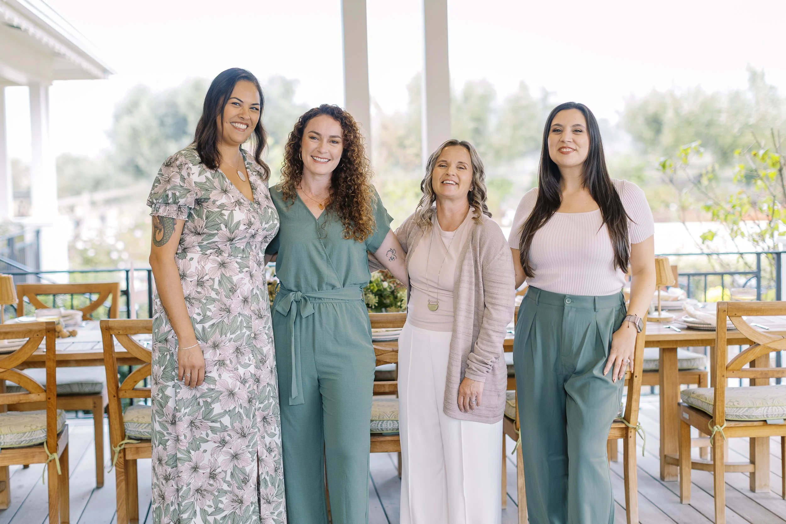 Four women standing together on a balcony, smiling at the camera, with a dining area in the background and outdoors visible beyond.