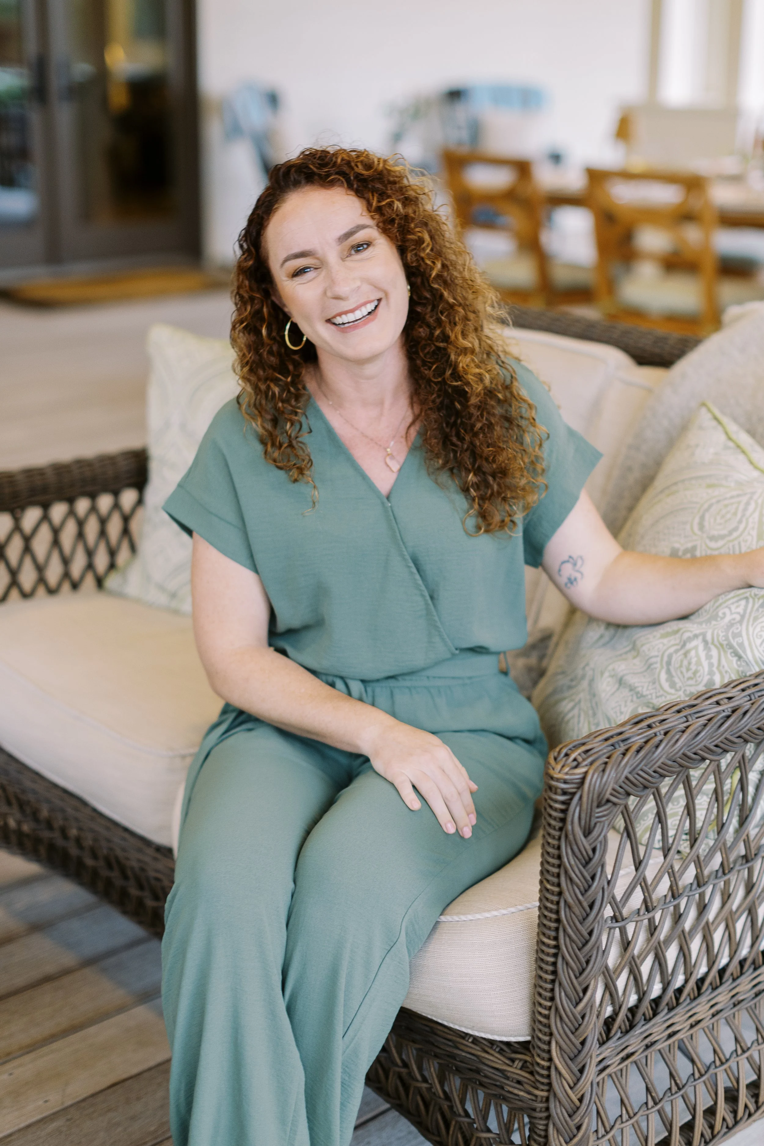 A woman with curly brown hair smiling outdoors on a sunny day, wearing a light blue cropped blouse, jeans, a pink pendant necklace, and beaded bracelets.