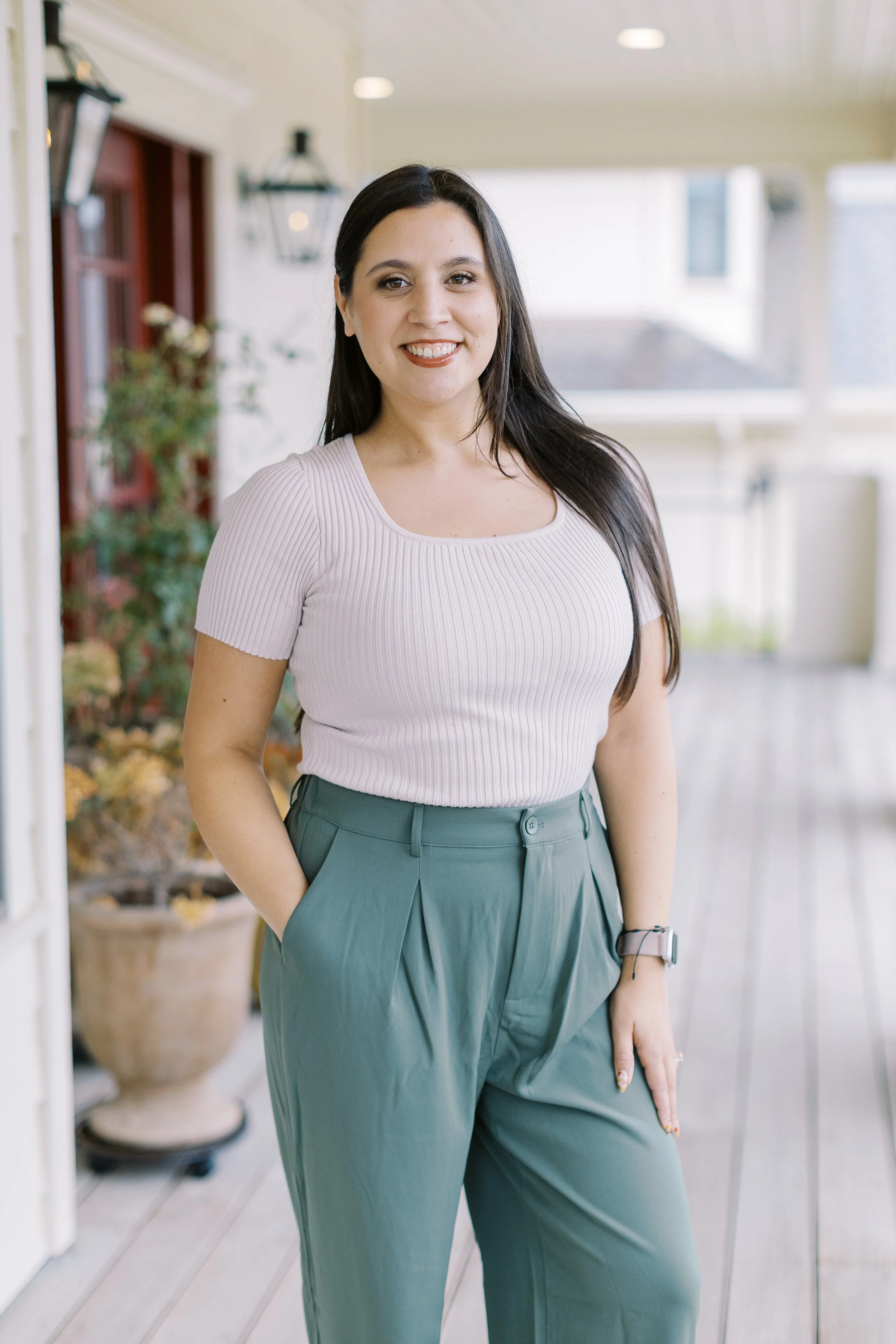 A woman standing on a porch, smiling at the camera, wearing a light pink ribbed T-shirt and green high-waisted pants.