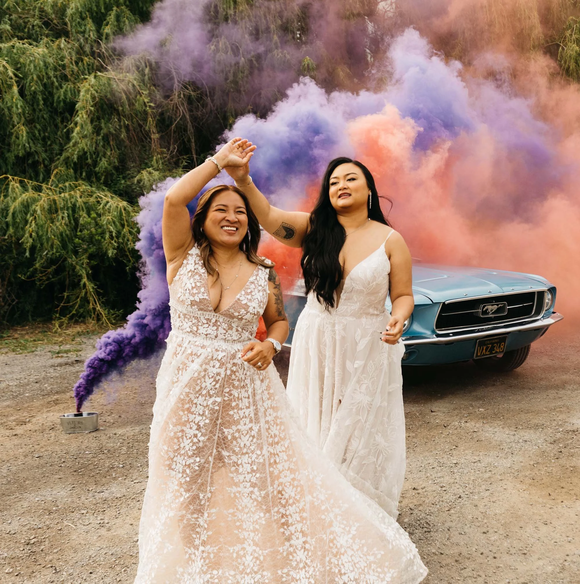 Two women in white dresses dancing and smiling in front of a blue vintage car with purple and pink smoke in the background.