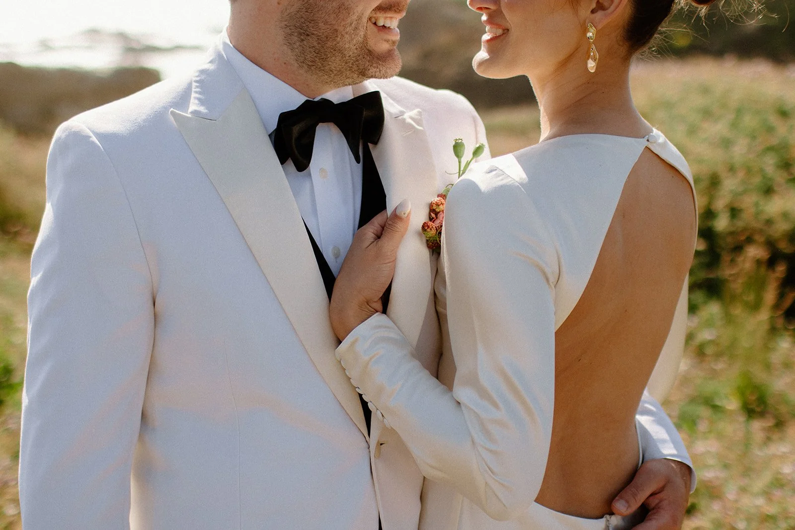 A bride and groom dressed in white wedding attire, standing close together outdoors, smiling and gazing at each other.