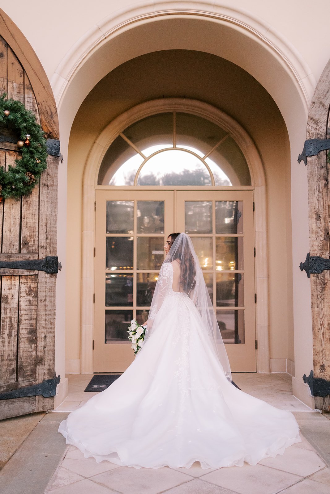 Bride in white wedding dress holding bouquet standing inside a building near a glass door with sunlight outside