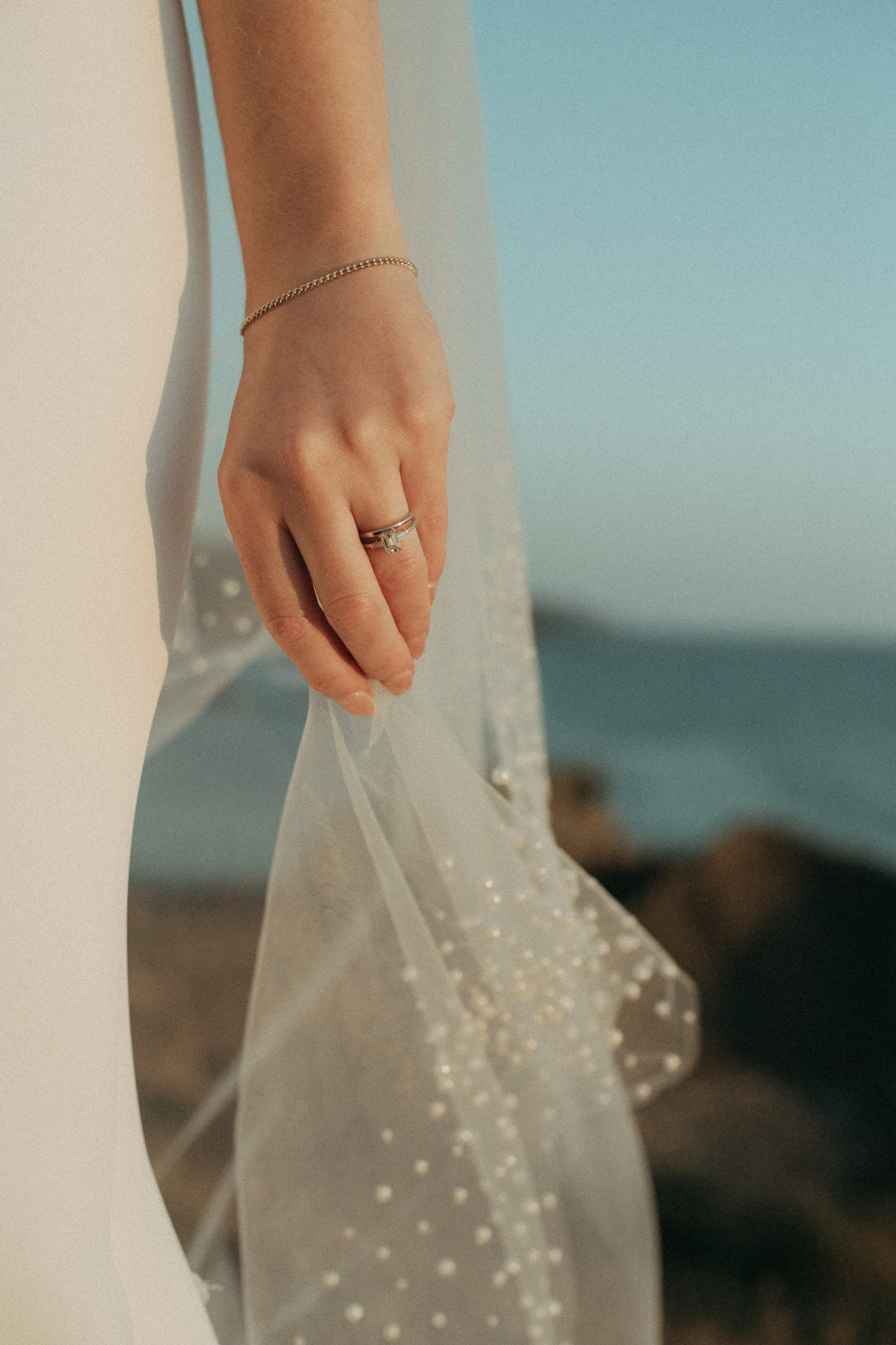 Close-up of a woman's hand with a ring and a bracelet, holding a sheer, pearl-embellished fabric against a beach background.