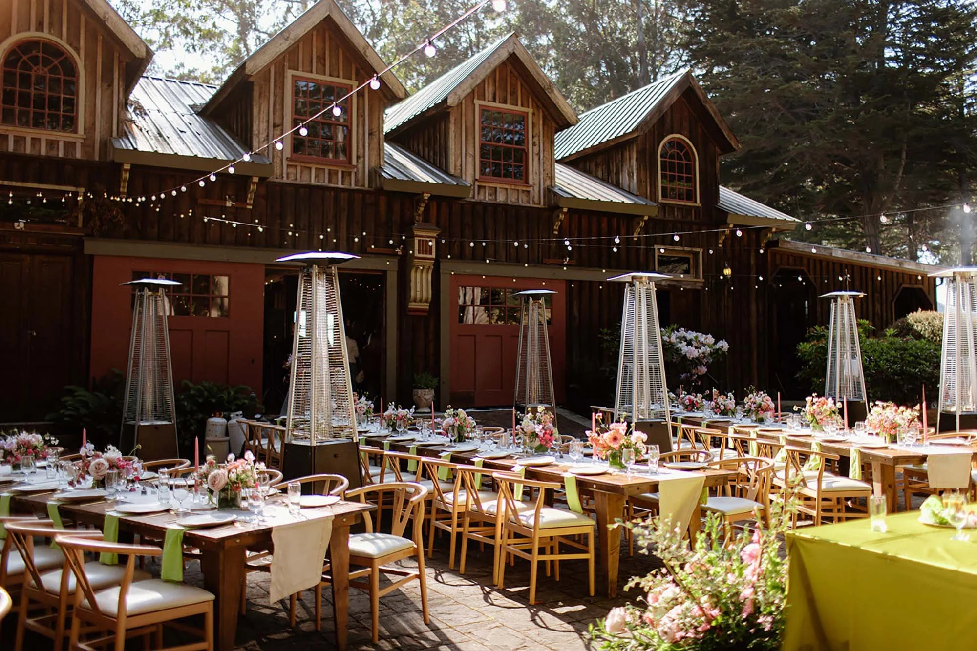 Outdoor event setup with wooden tables and chairs, decorated with floral arrangements, string lights, and tall outdoor heaters in front of a rustic wooden building with triangle-shaped roofs.