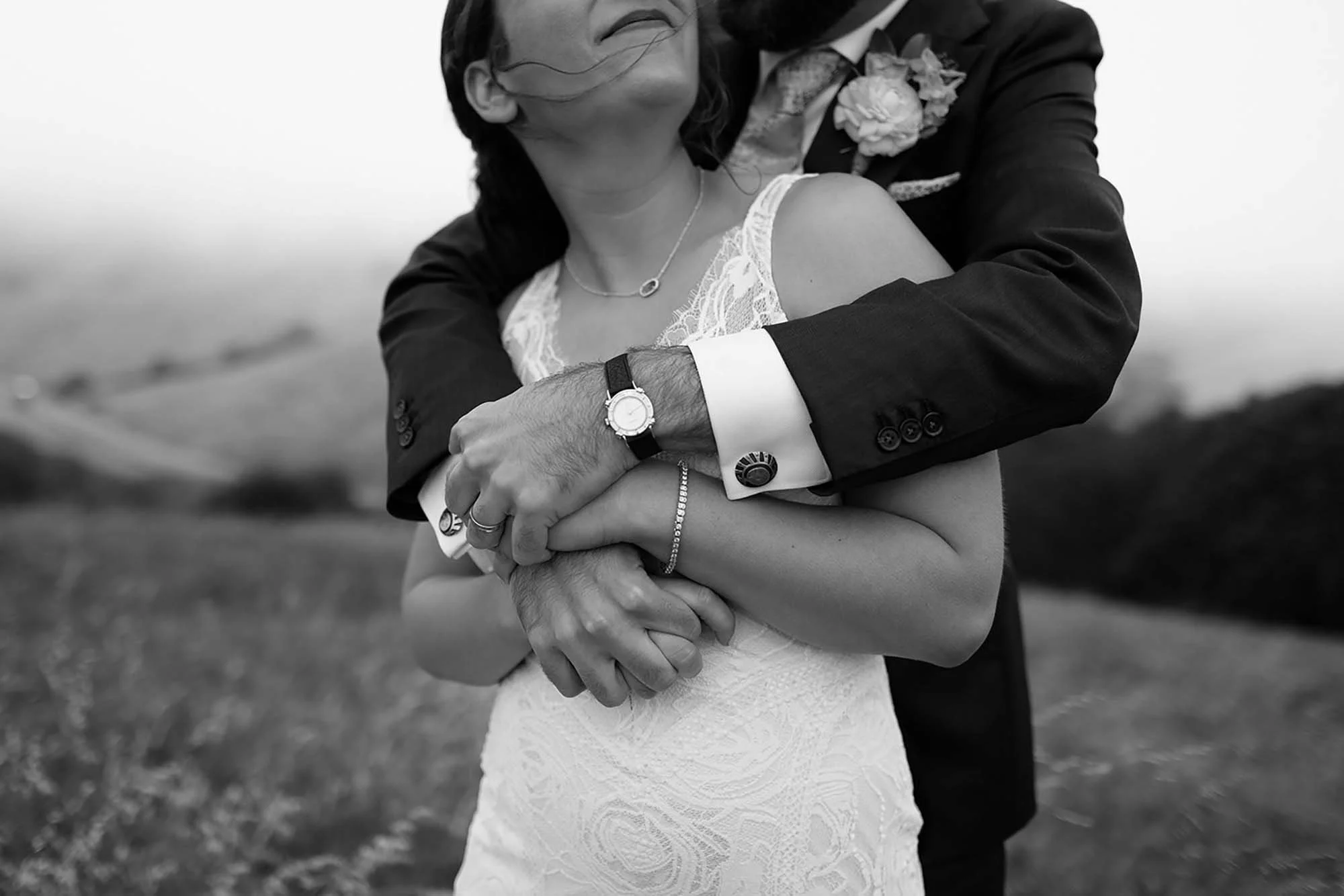 Black and white photo of a couple embracing outdoors, with the woman wearing a dress and the man in a suit. The focus is on their intertwined hands and arms, showing wedding rings, watches, and jewelry, with a blurred landscape in the background.