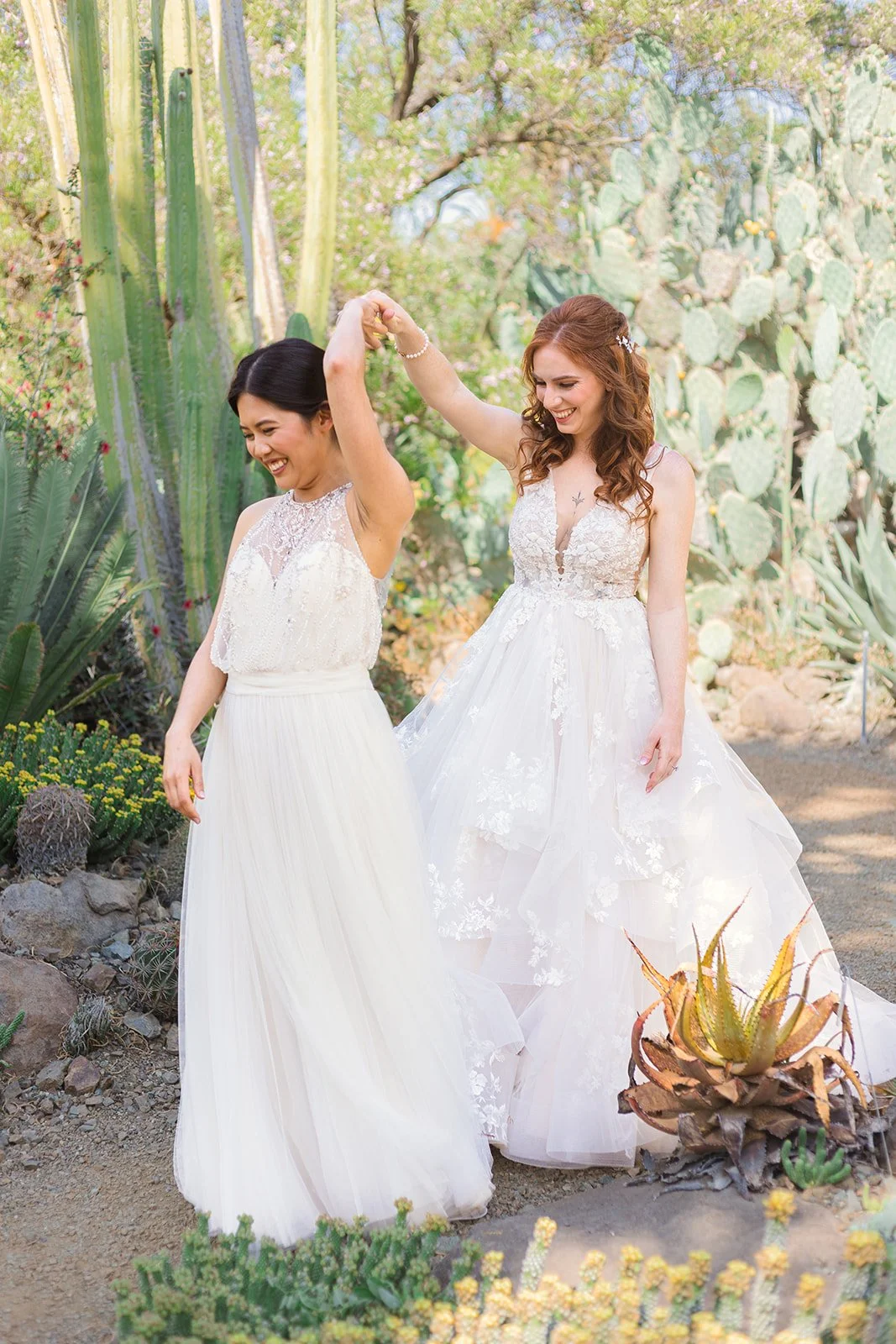 Two women in wedding dresses dancing in a desert garden with cacti and succulents, smiling and enjoying themselves.