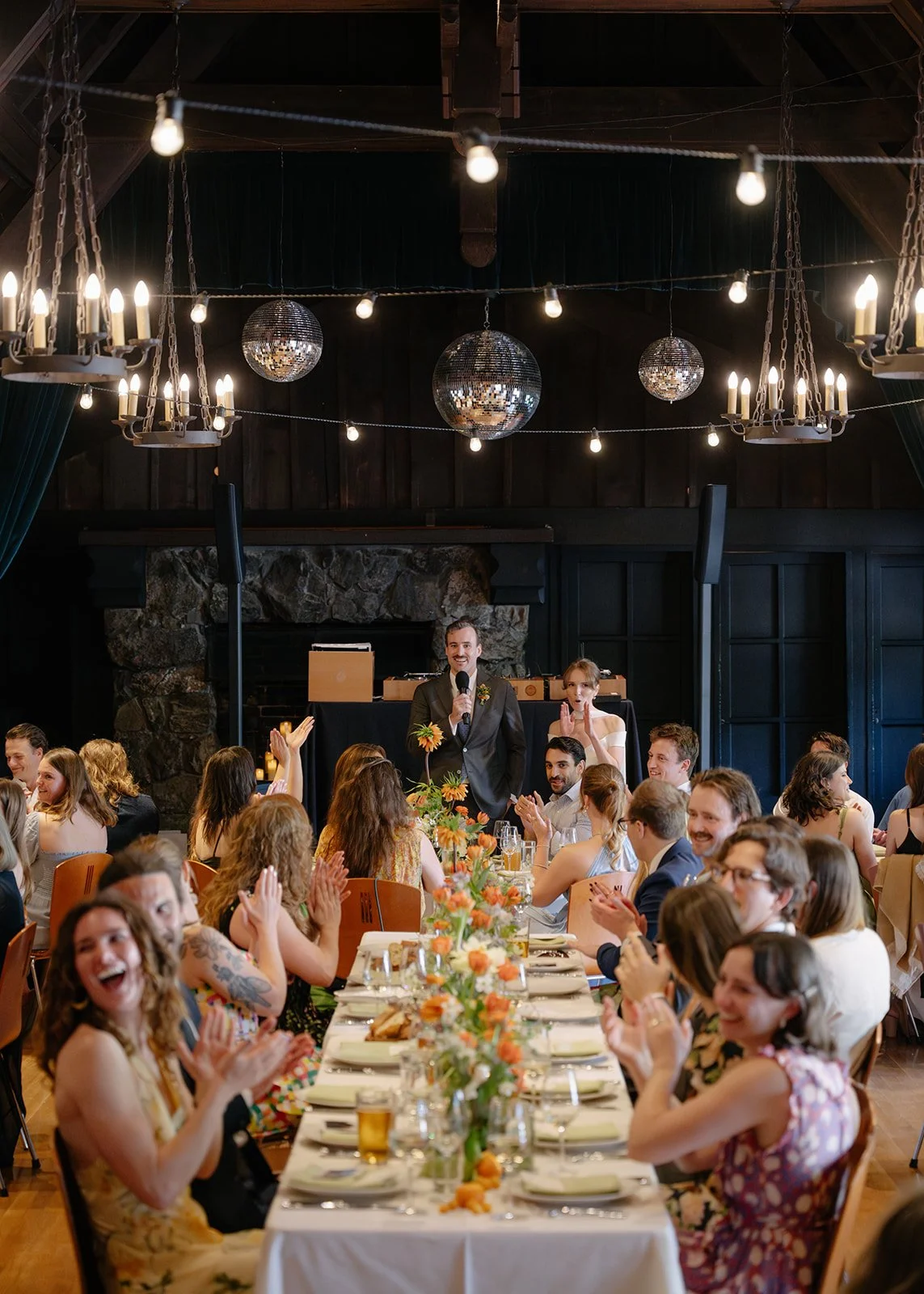 People attending a wedding reception, with a bride and groom. The groom is at a microphone, smiling, while guests are clapping and laughing around a decorated banquet table, in a rustic venue with chandeliers and disco balls.
