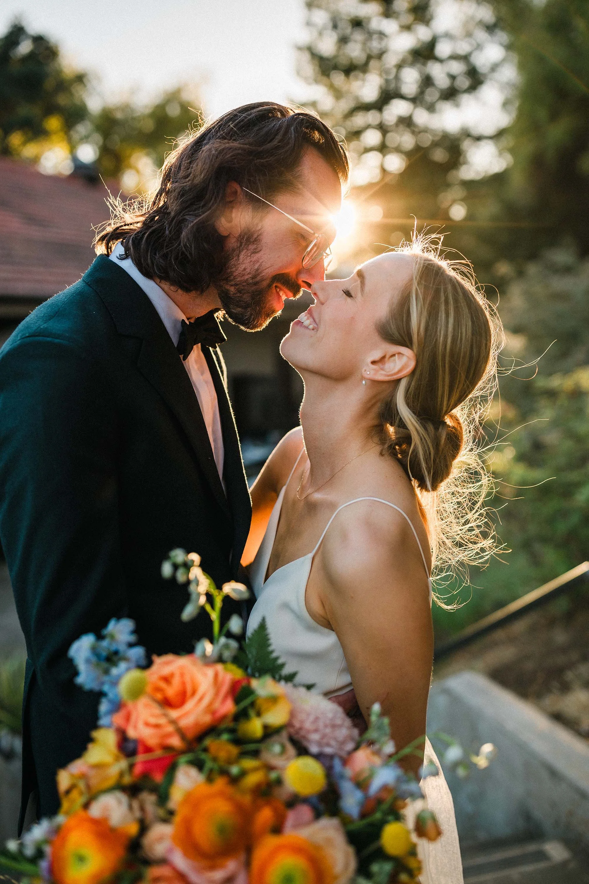 A couple at their wedding during sunset, smiling and leaning towards each other, with the bride holding a colorful bouquet of flowers.
