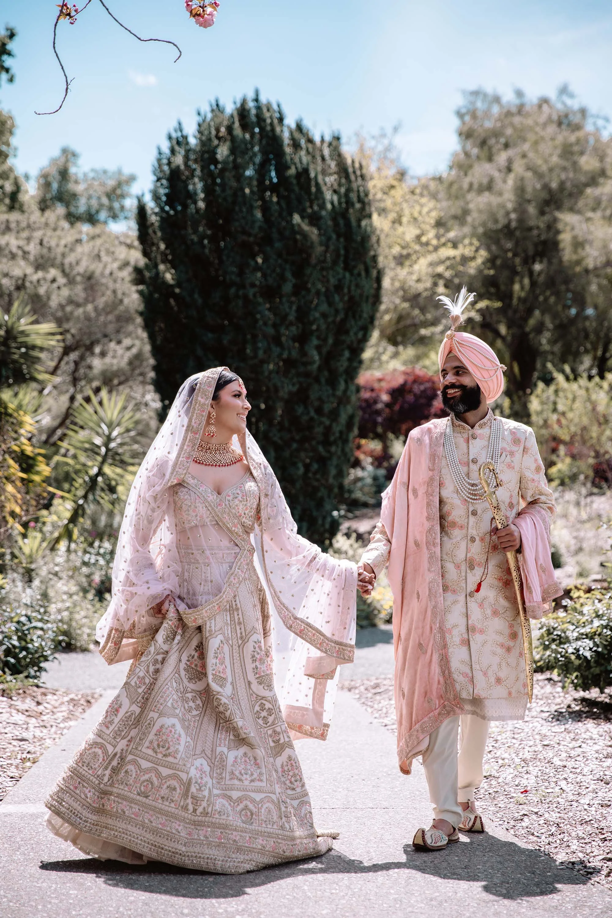 A couple in traditional Indian wedding attire holding hands and smiling at each other outdoors in a garden setting.