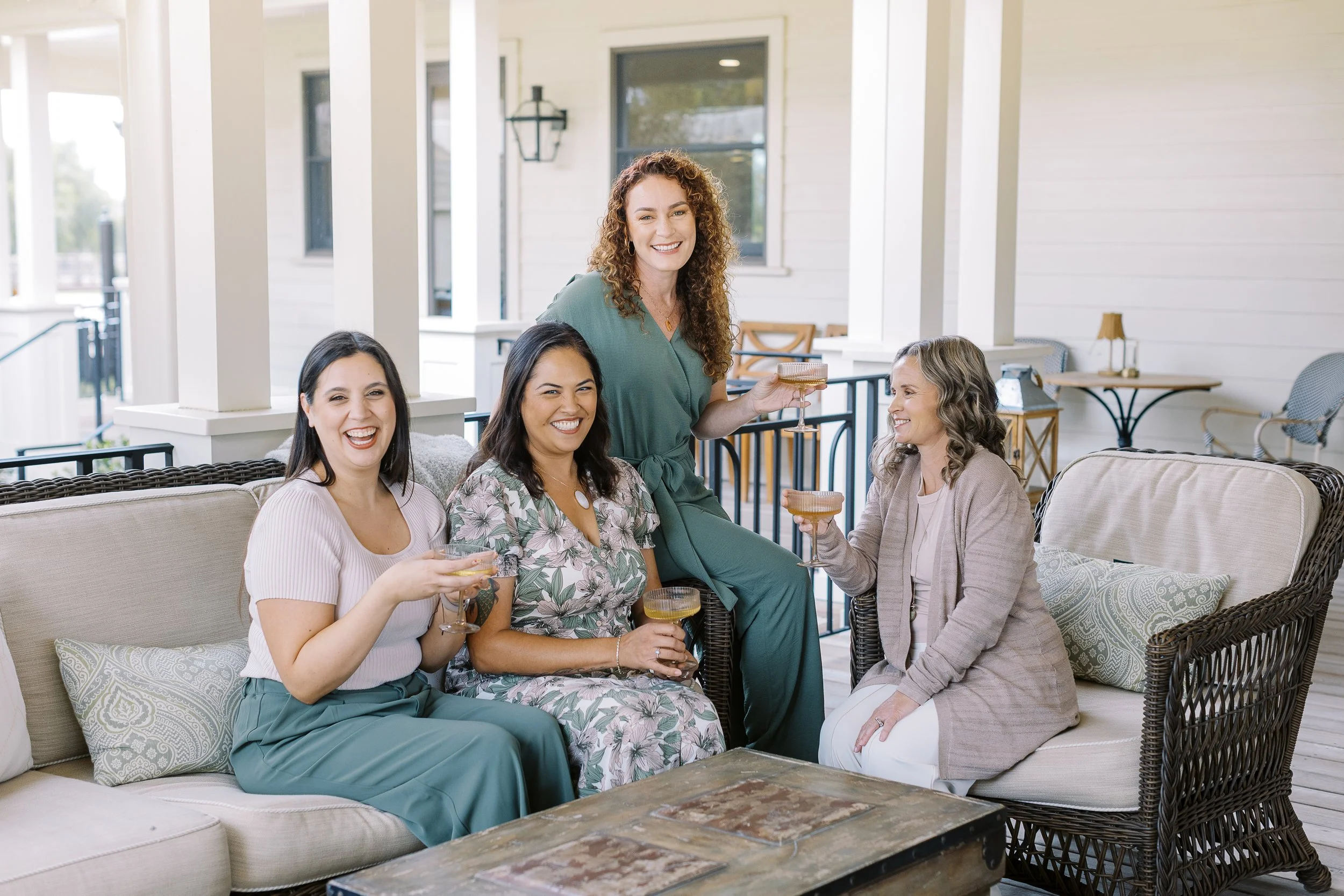 Four women sitting and standing on a porch, holding cocktails, smiling and enjoying each other's company.