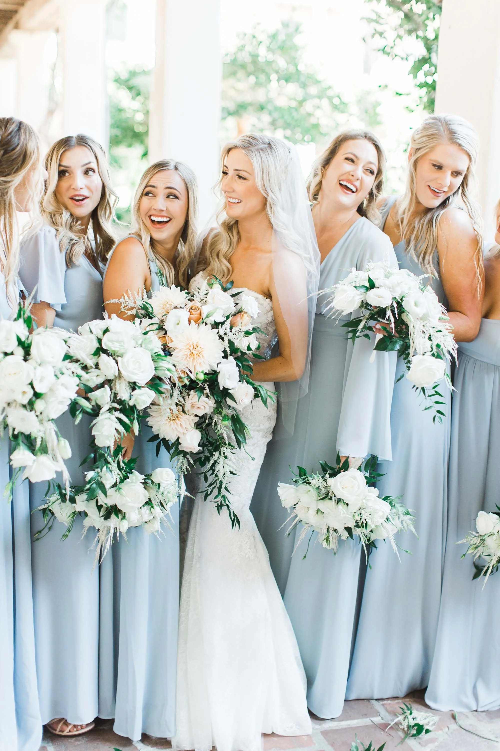 Bride in white wedding gown surrounded by bridesmaids in pale blue dresses, all holding white and blush flower bouquets at a wedding celebration.