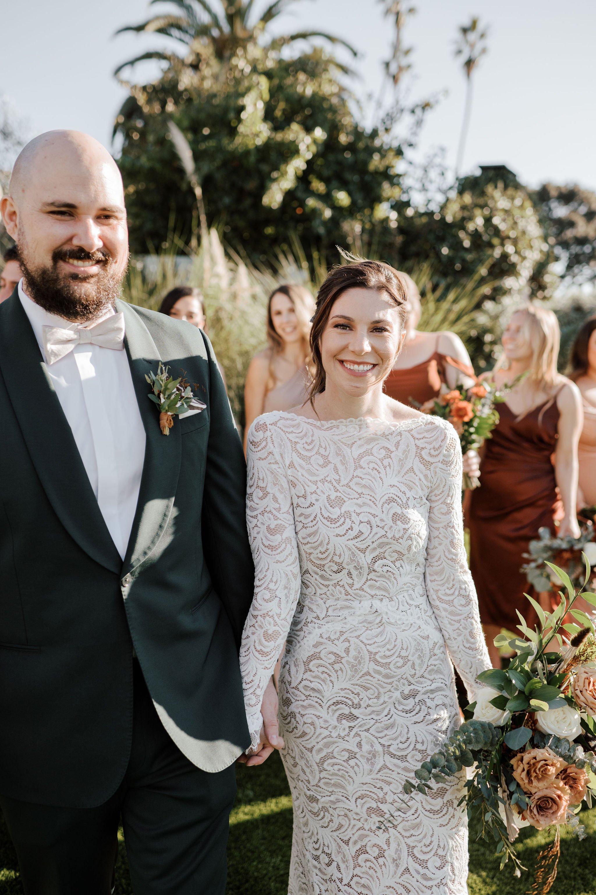 A bride and groom holding hands at their outdoor wedding ceremony, smiling with friends in the background, greenery, and sunlight.