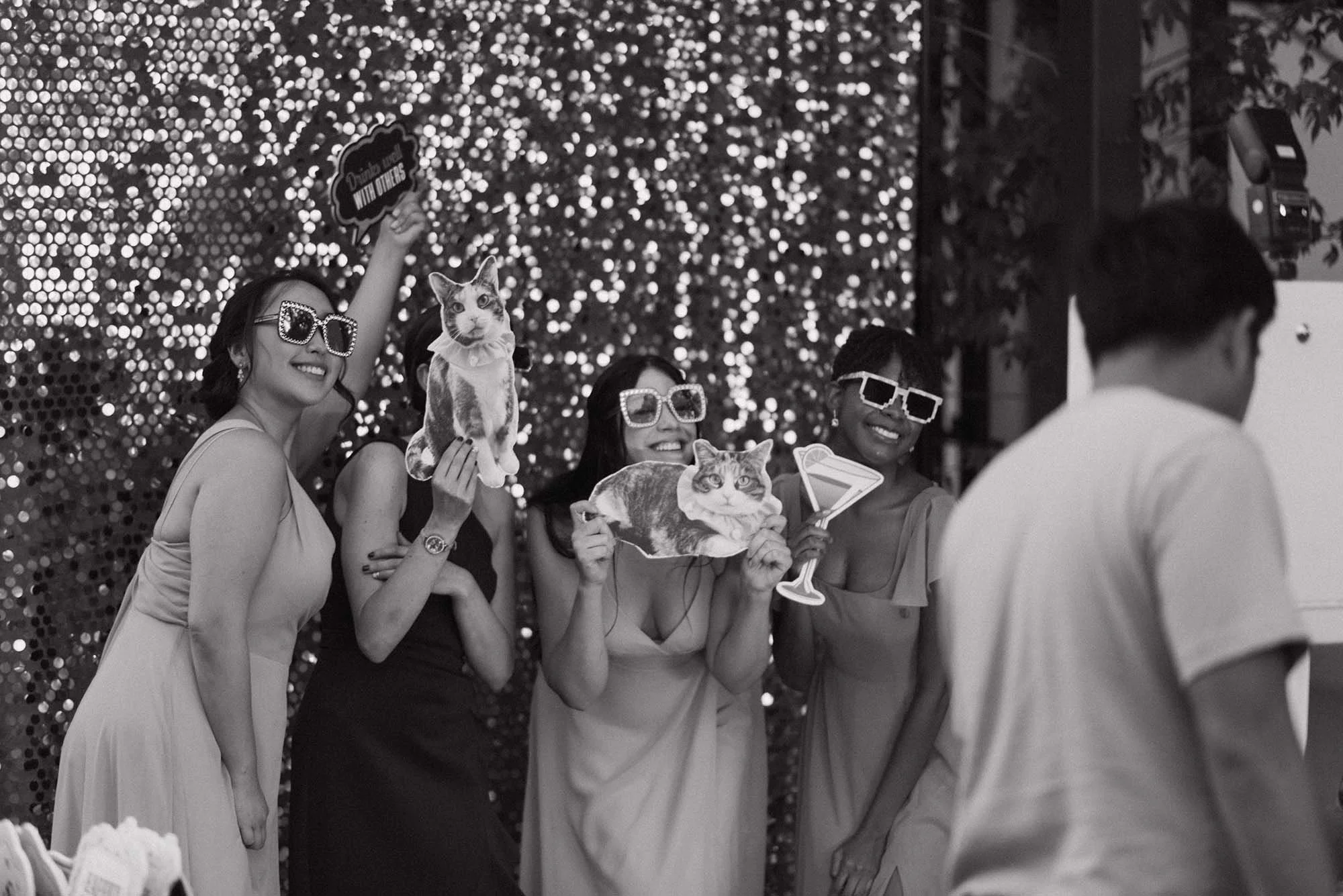 Four women at a party with photo props, including cats and drinks, smiling and posing for the camera. One woman is holding a photo of a cat, and another is holding a paper cutout of a martini glass. They are standing in front of a sparkly backdrop, wearing sunglasses, with a man in the foreground.