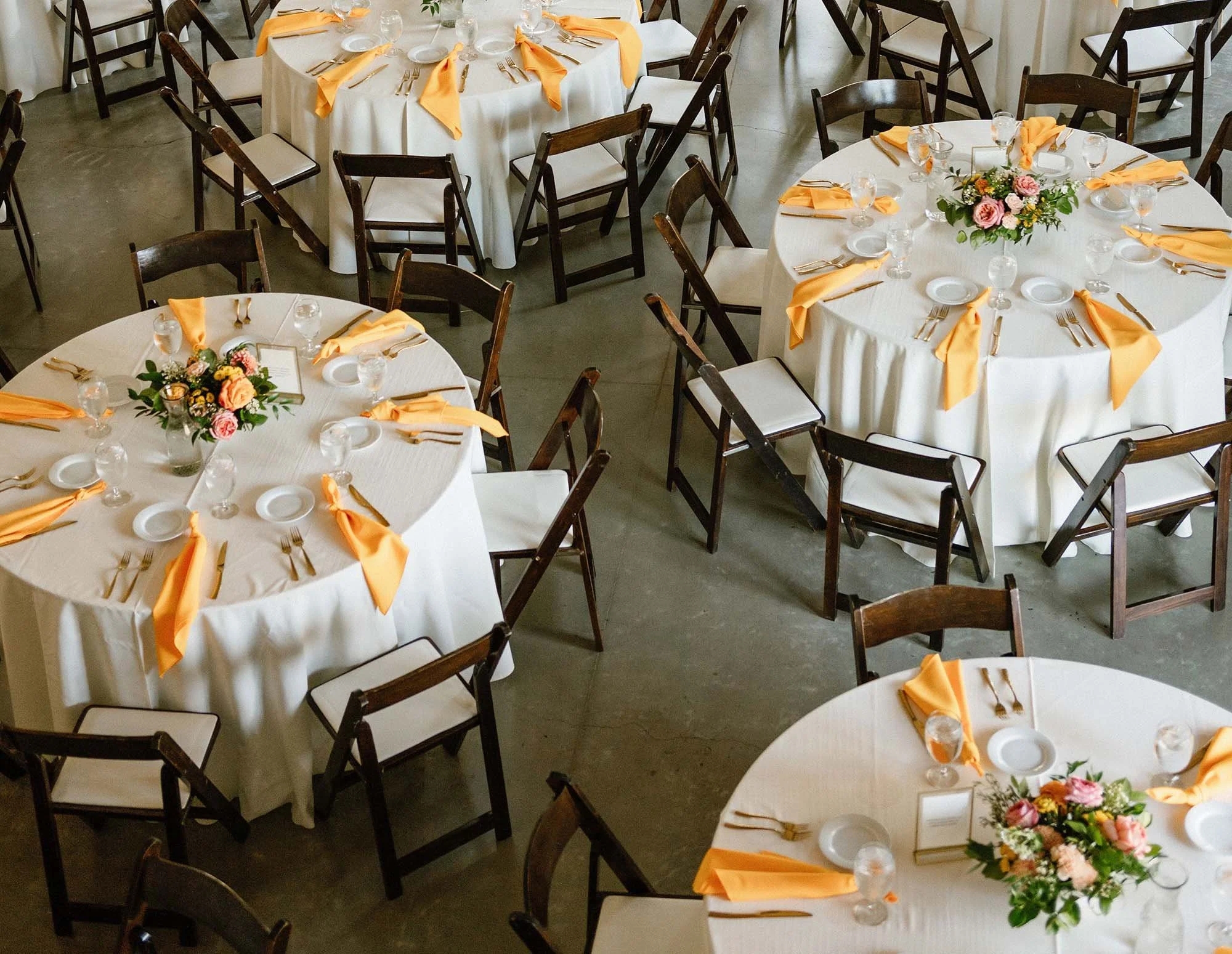 Round banquet tables covered with white tablecloths, decorated with floral centerpieces, orange napkins, white plates, gold utensils, and water glasses. Brown chairs surround the tables, set for a formal event.