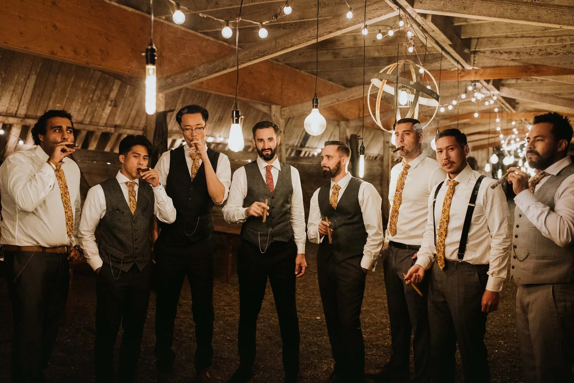 A group of nine men dressed in vintage-style formal clothing, including vests, suspenders, and ties, are standing together in a rustic, wooden barn-like setting decorated with hanging string lights and Edison bulbs.