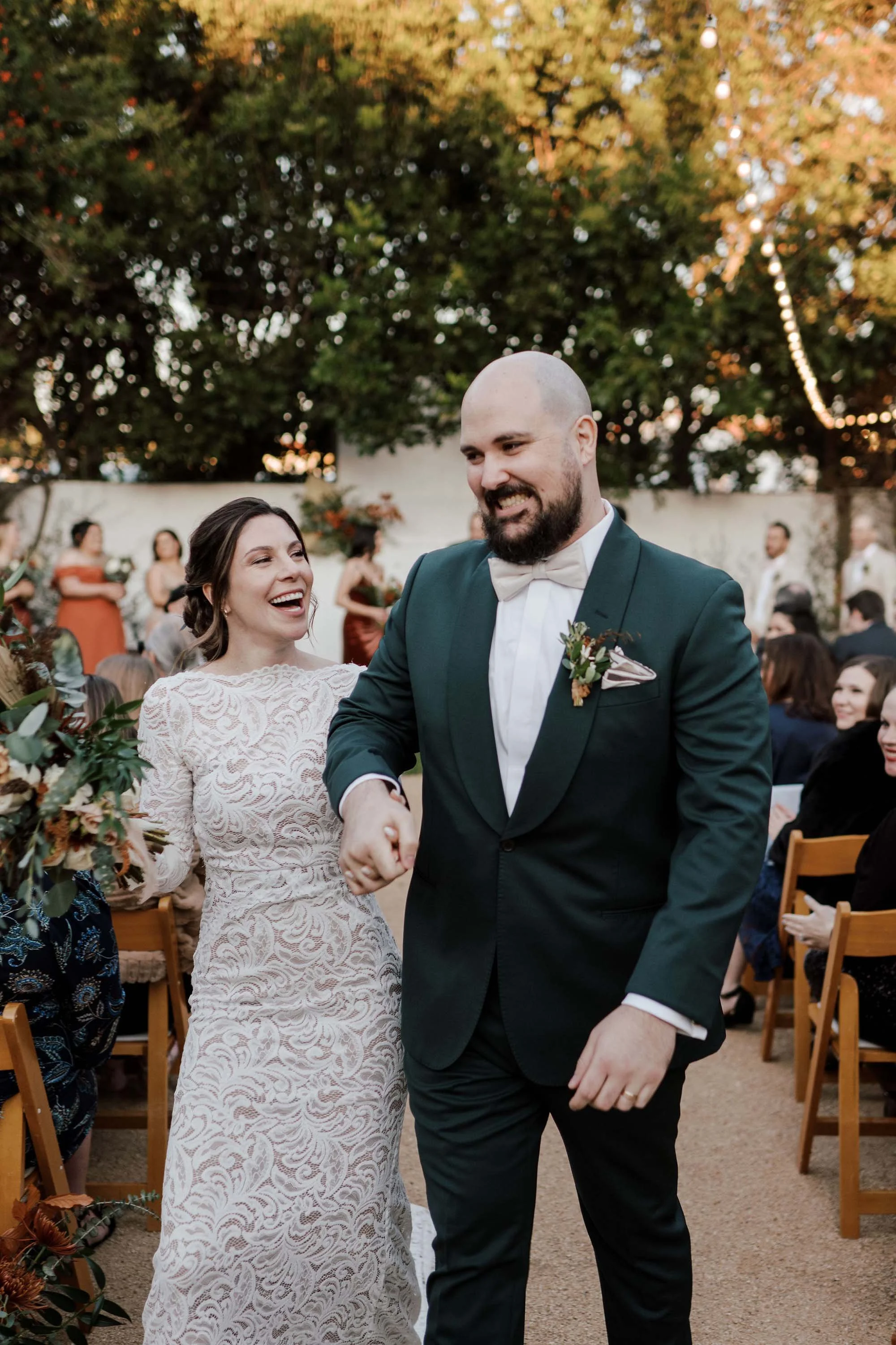A bride and groom holding hands and smiling at each other during their outdoor wedding ceremony, surrounded by guests and decorated with greenery and flowers, with string lights hanging in the background.