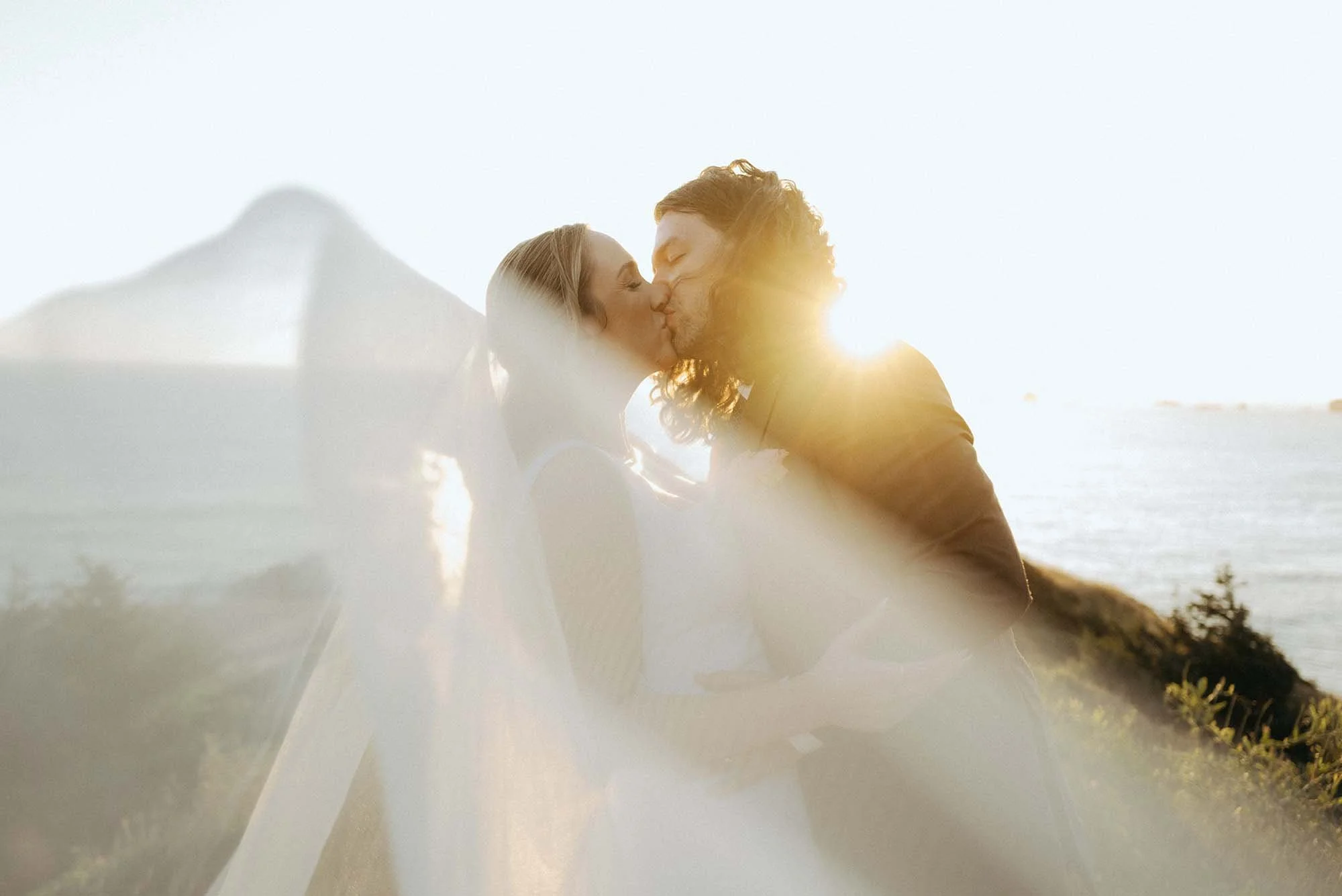 A couple kissing at sunset in an outdoor setting with water in the background.