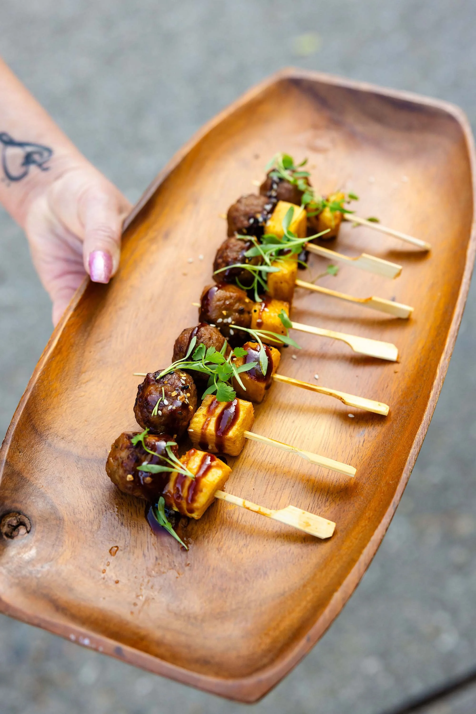Skewers with grilled meat and pineapple garnished with microgreens on a wooden serving tray.
