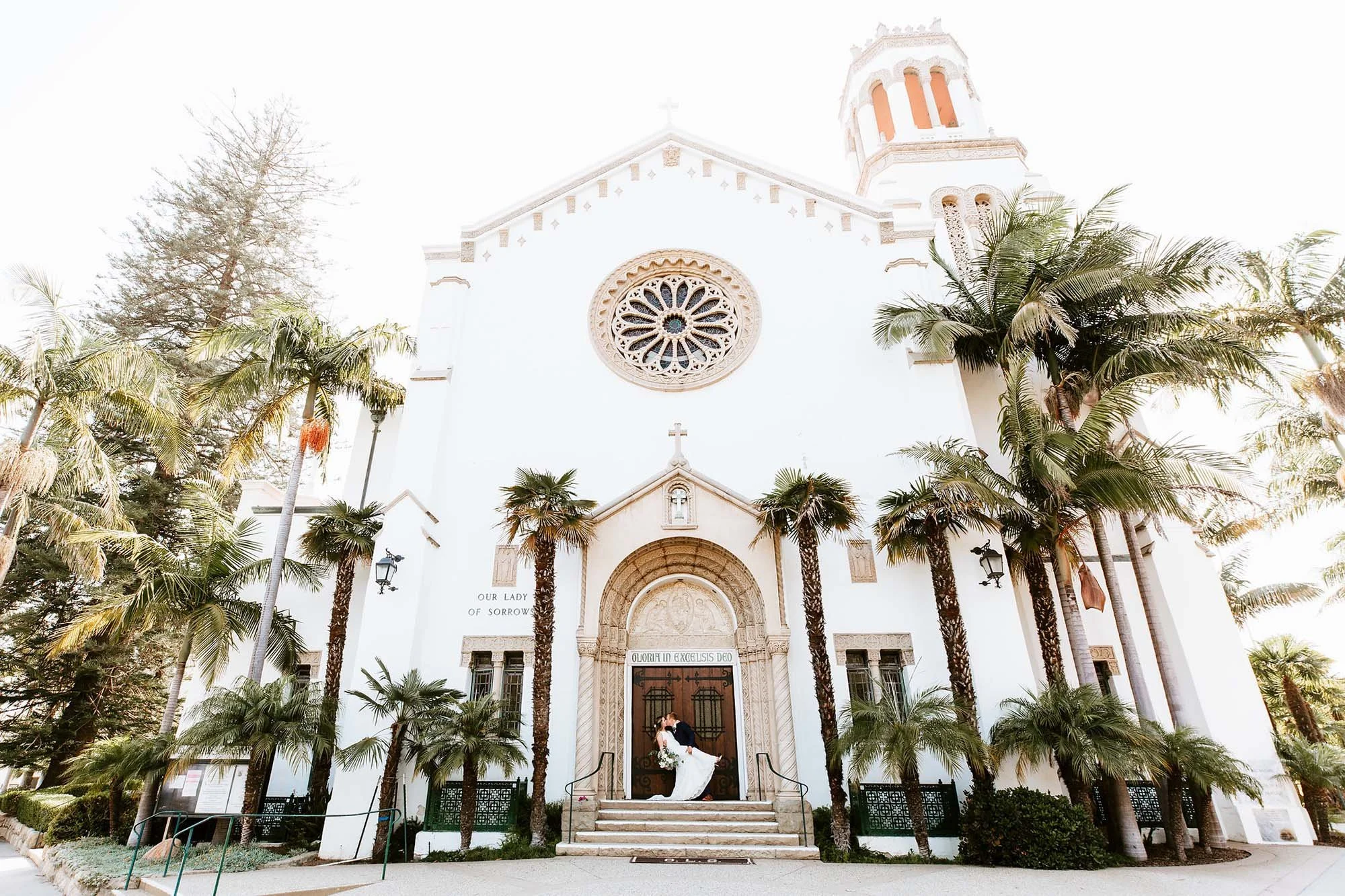 Bride and groom standing in front of a white church with palm trees and stone stairs.