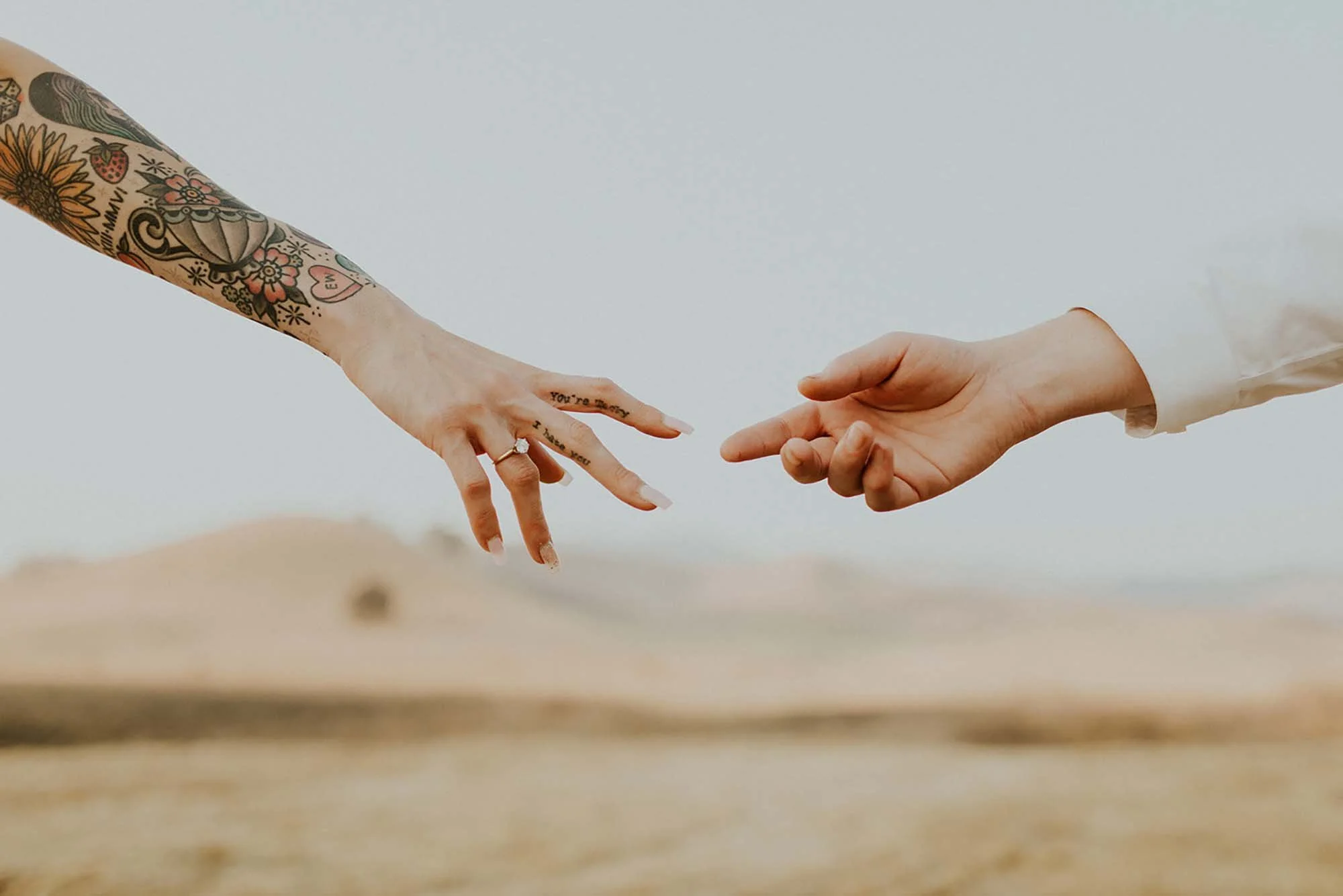 Two hands reaching out to each other, with one hand decorated with colorful tattoos and the other hand plain, against a blurred beach background.