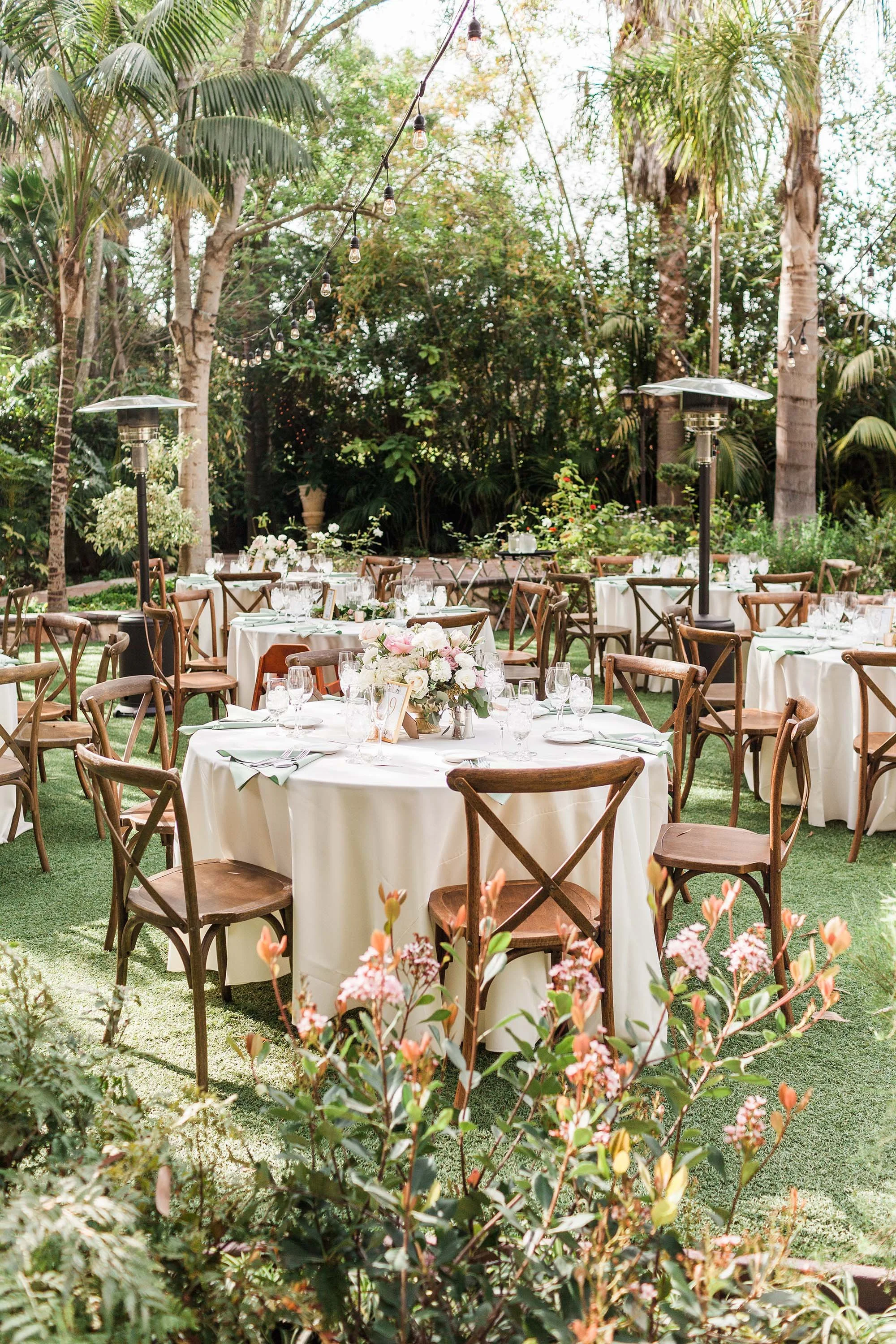 Outdoor garden wedding reception setup with round tables, white tablecloths, floral centerpieces, wooden chairs, string lights, and tall patio heaters amidst trees and greenery.