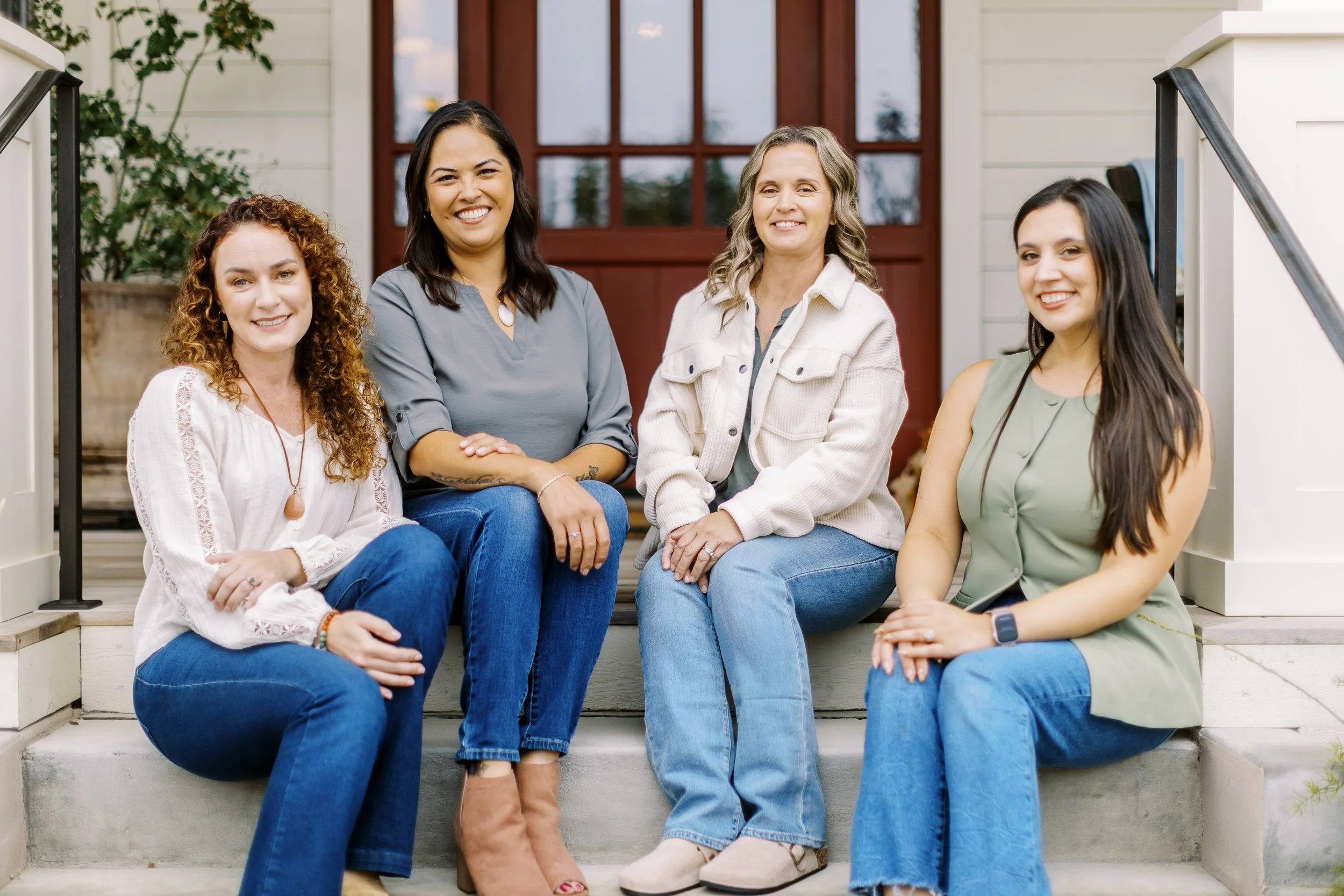 Four women sitting on the steps of a house, smiling at the camera. The house has a red door and white siding.