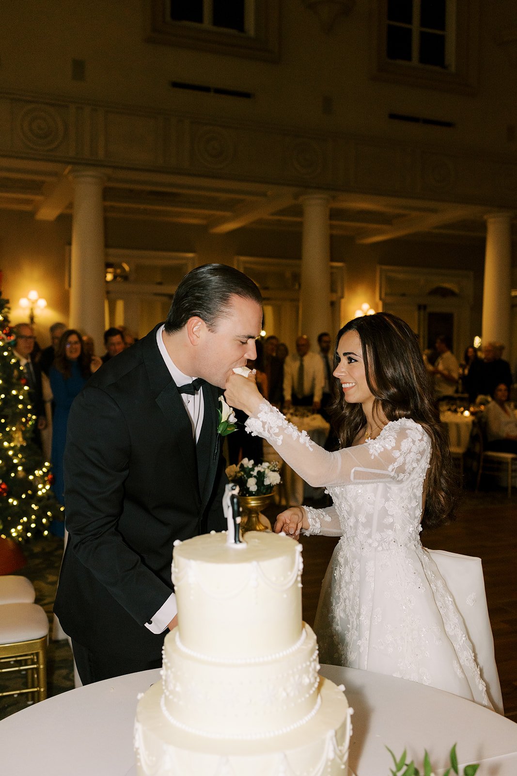 A bride feeds cake to a groom at their wedding reception, with guests in the background and a decorated Christmas tree.