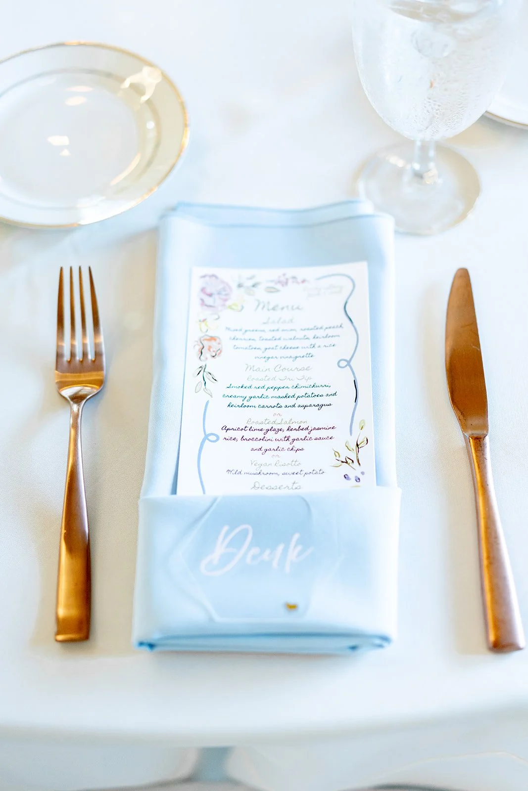 Elegant table setting with a light blue napkin holder holding a menu, a water glass, a gold fork on the left, and a gold knife on the right, on a white tablecloth.