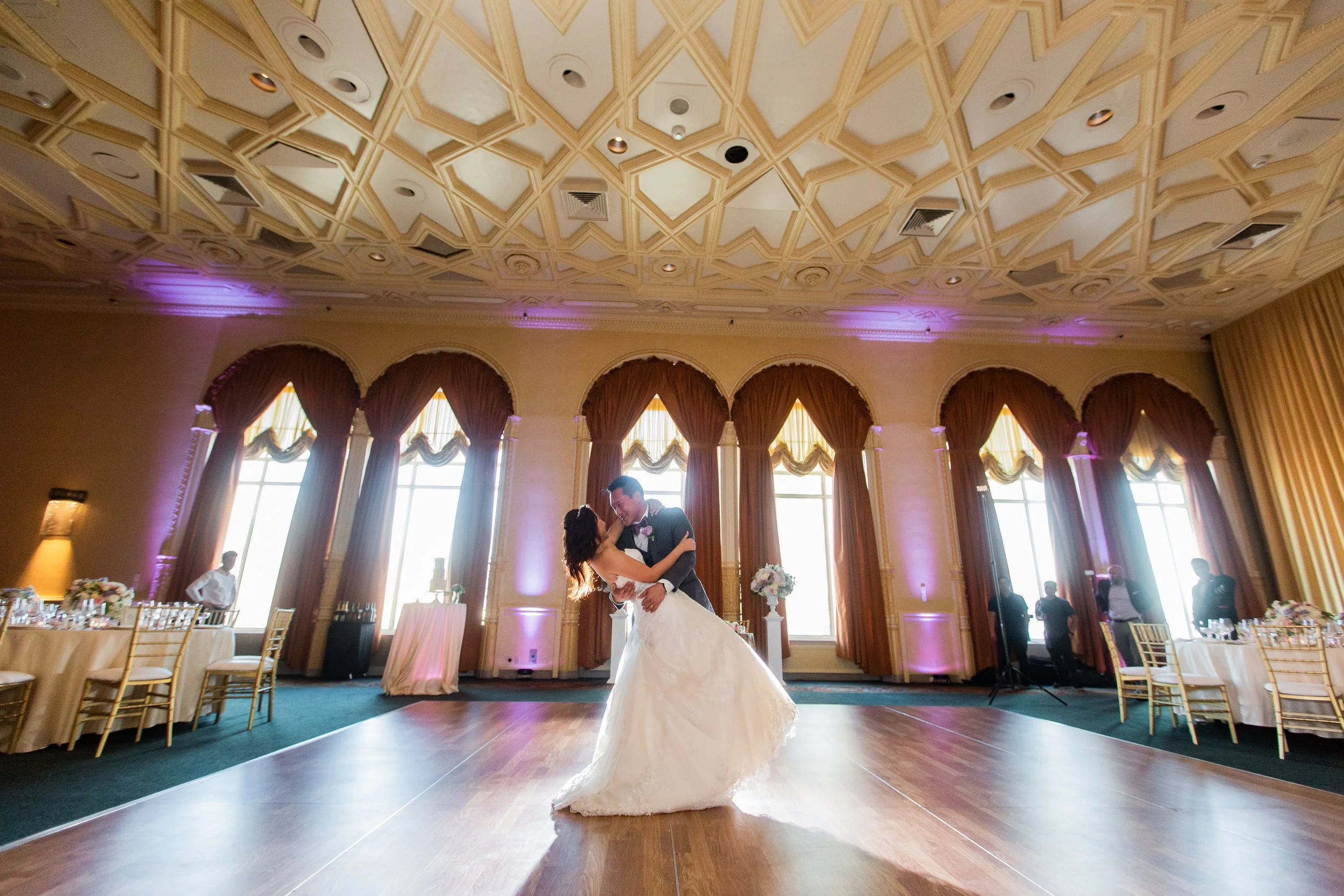A bride and groom dance together in a grand, elegant wedding reception hall with large windows and draped curtains.