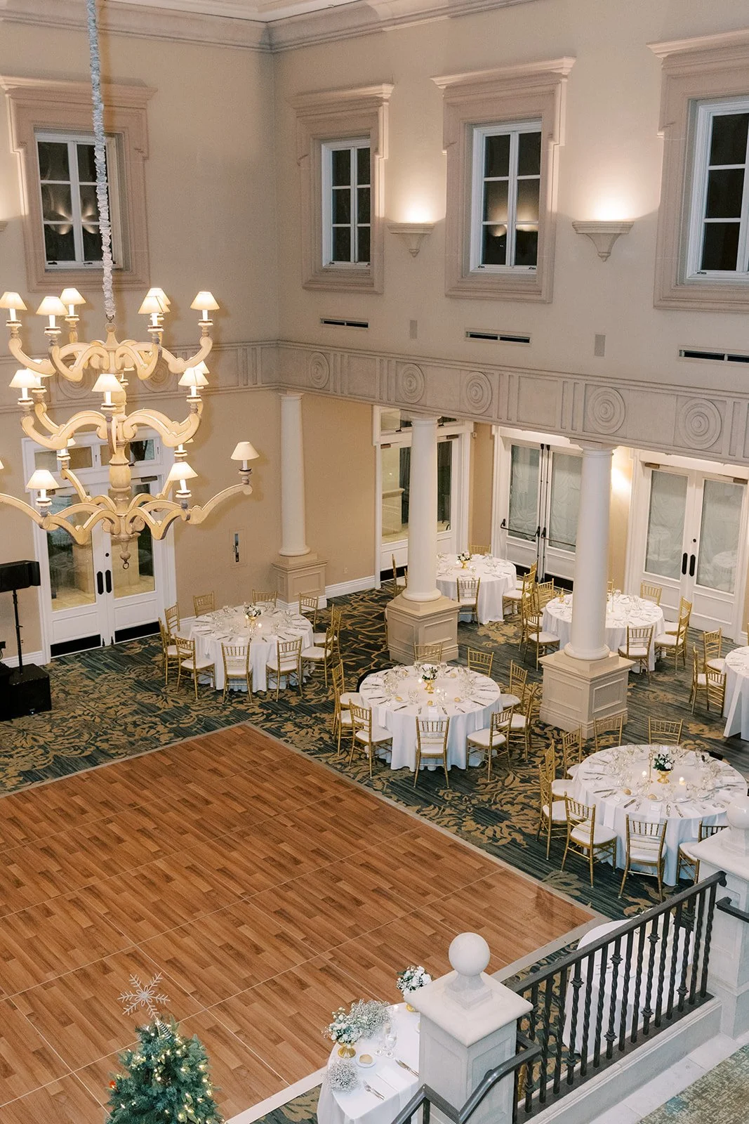 Elegant banquet hall with round tables draped in white tablecloths, surrounded by gold chairs, and a wooden dance floor. A chandelier hangs from the ceiling, and the room has tall windows with white trim, cream walls, and decorative columns.