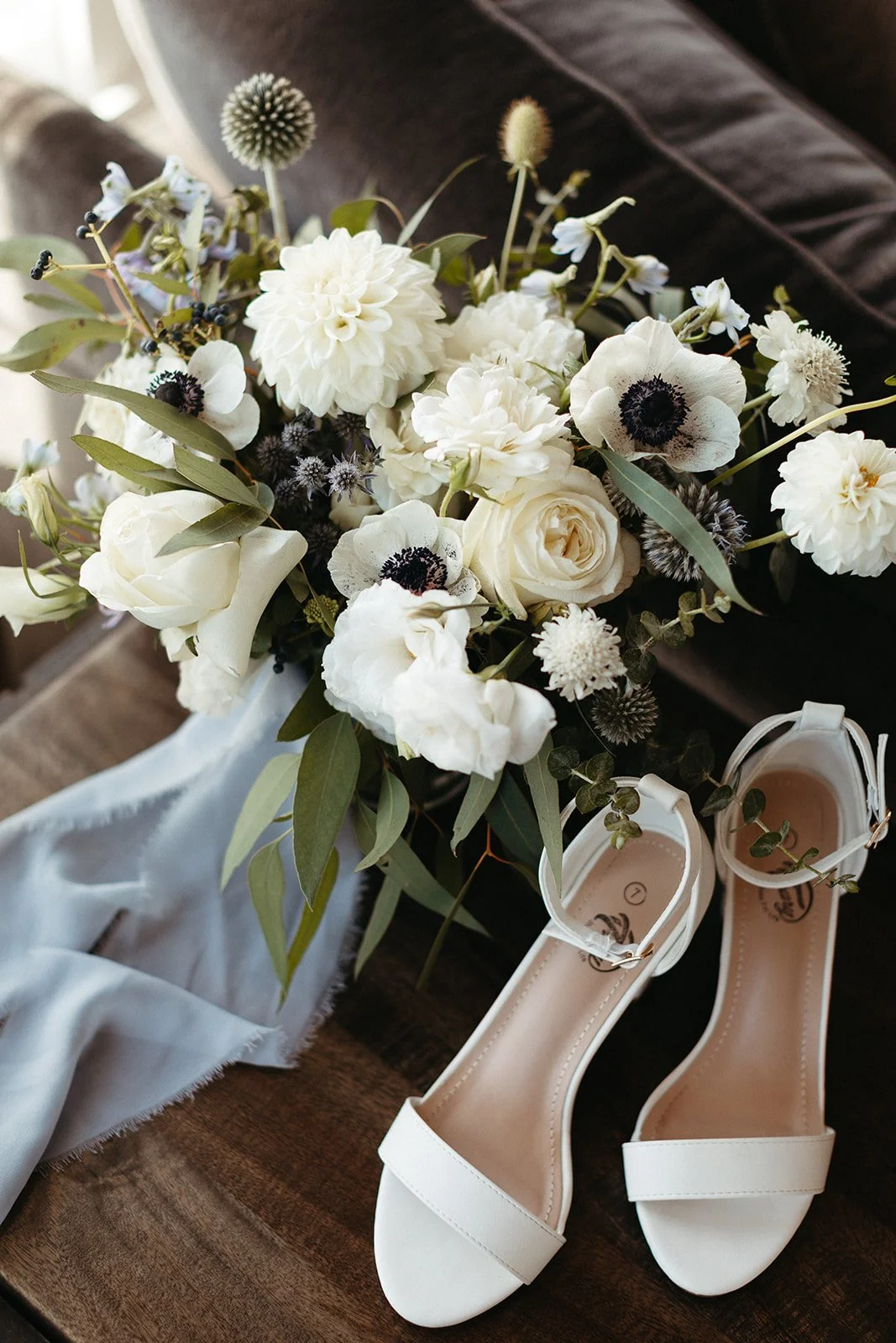 A bouquet of white flowers, including roses, anemones, and dahlias, with green leaves, placed on a dark wooden surface next to a pair of white open-toe high-heeled shoes.