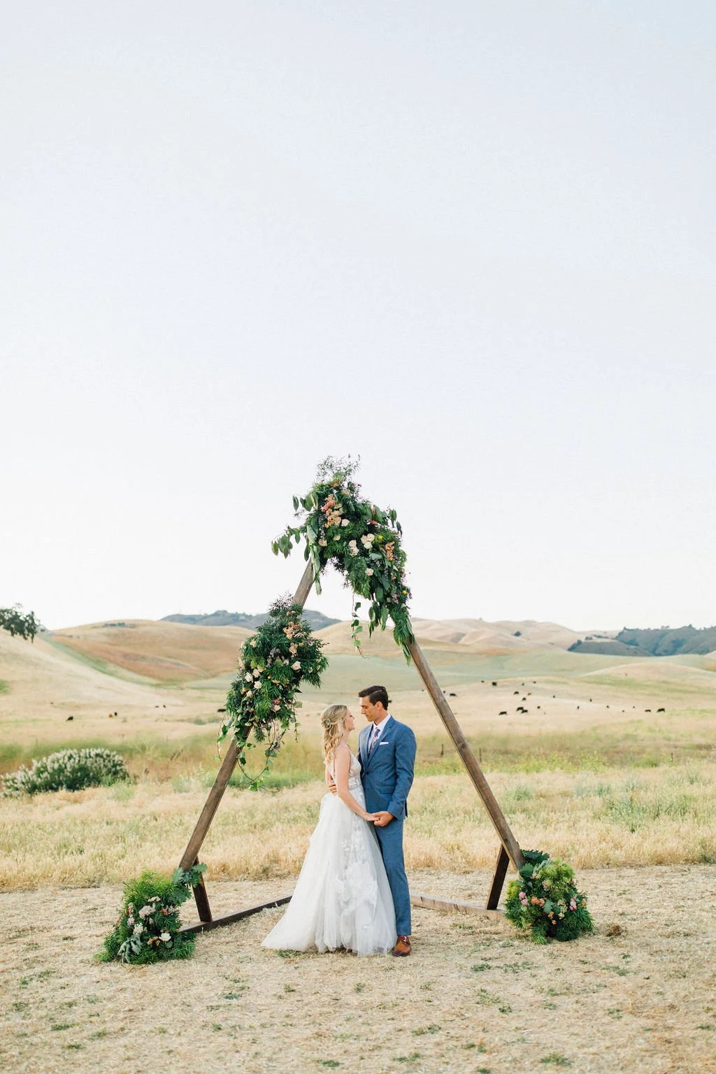 A bride and groom standing together under a wooden wedding arch decorated with flowers and greenery in an outdoor field with hills in the background.