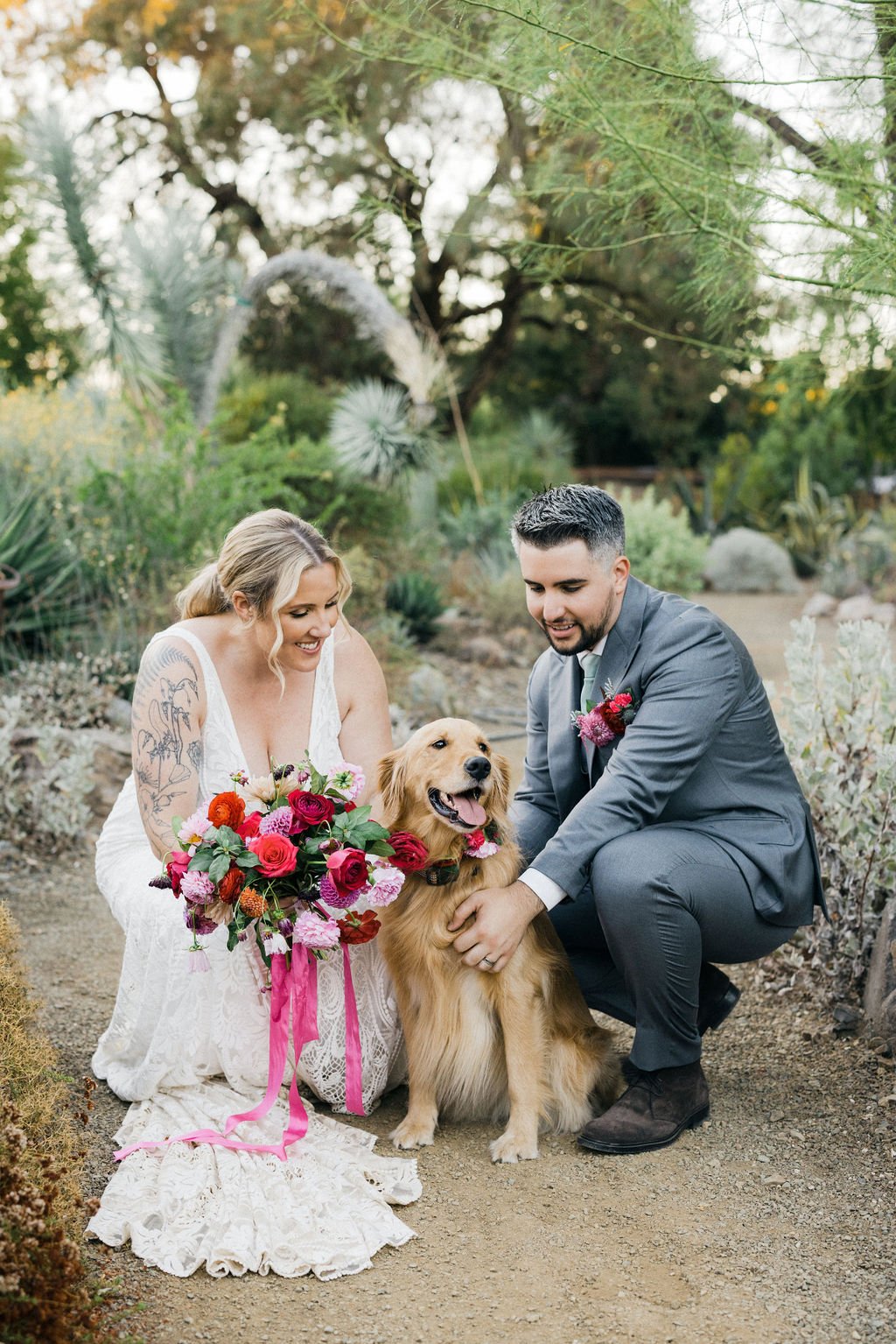 A bride and groom kneeling outdoors with a golden retriever dog, holding its head and collar decorated with flowers, surrounded by desert plants and trees.