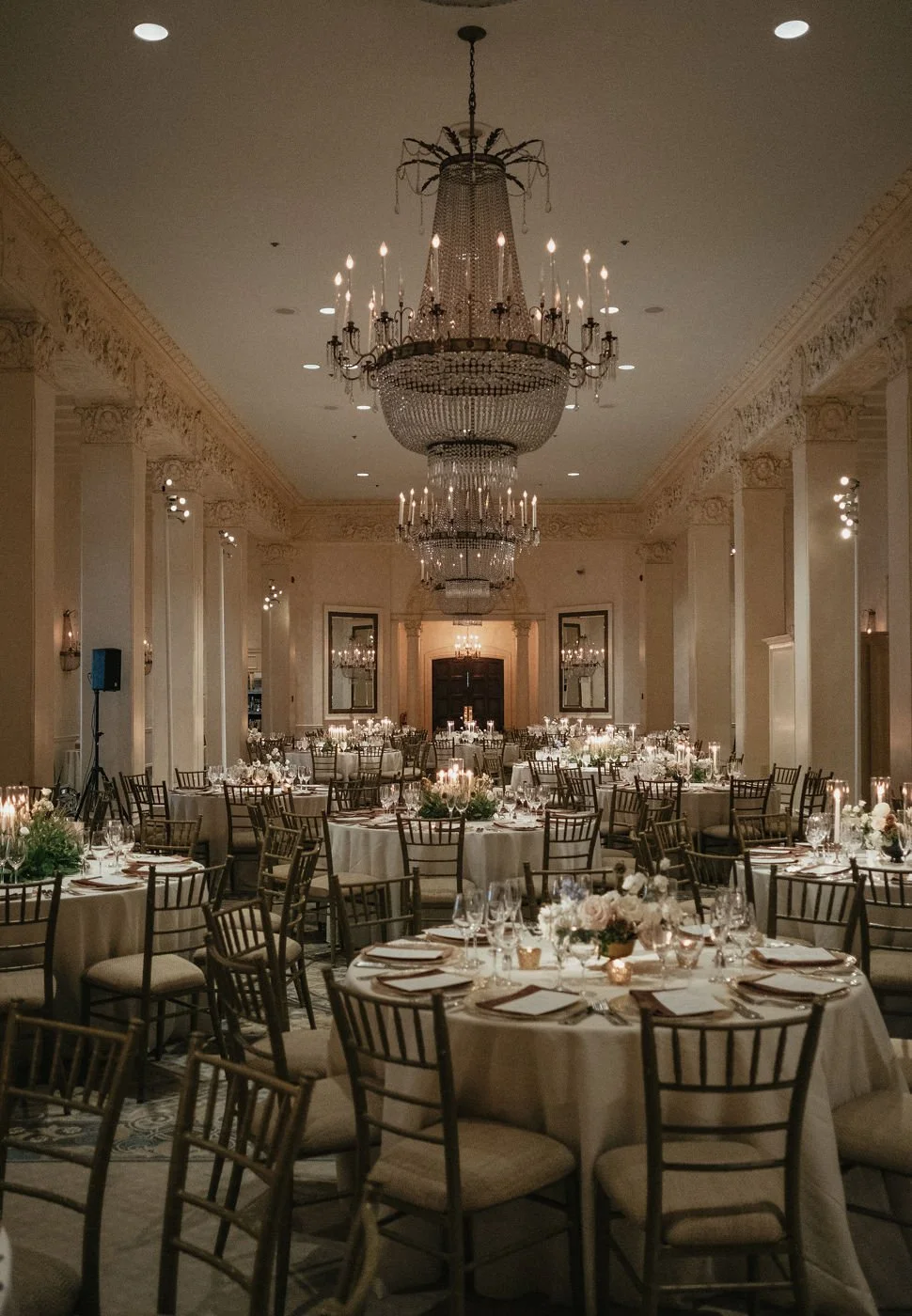 Elegant banquet hall decorated for a formal event with round tables covered in white tablecloths, floral centerpieces, and multiple chandeliers hanging from the ceiling.