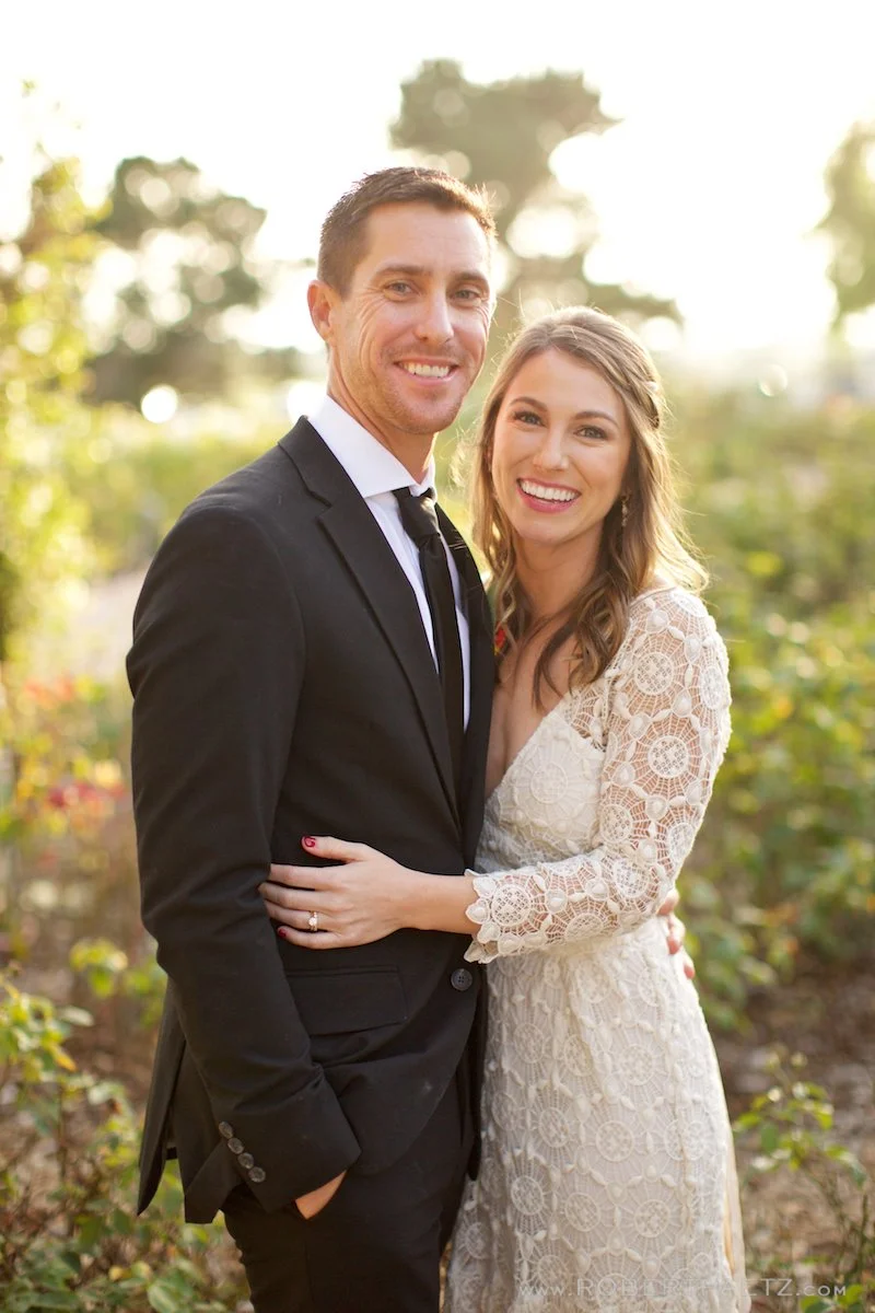 A smiling couple looks at the camera outdoors in a natural setting with trees and sunlight in the background. The man wears a dark suit and tie, and the woman wears a white lace dress with floral patterns.