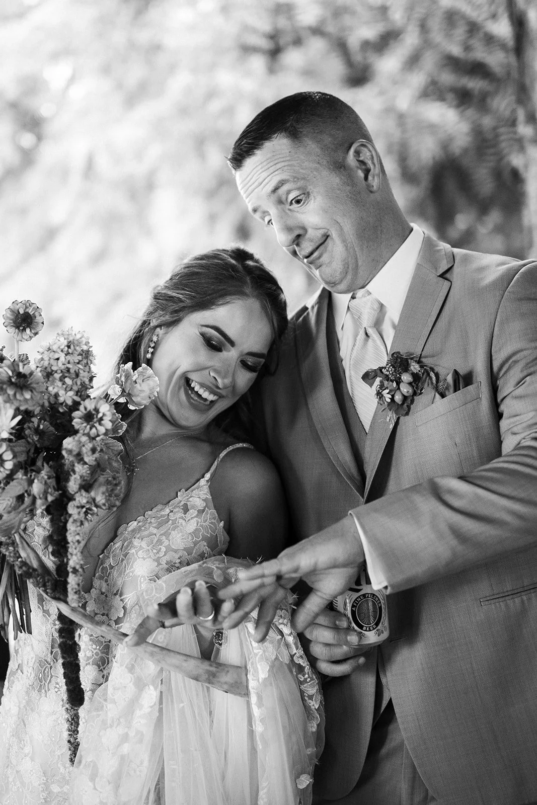 Black and white photo of a happy bride and groom looking at a phone together, the groom holding a beer, both smiling.