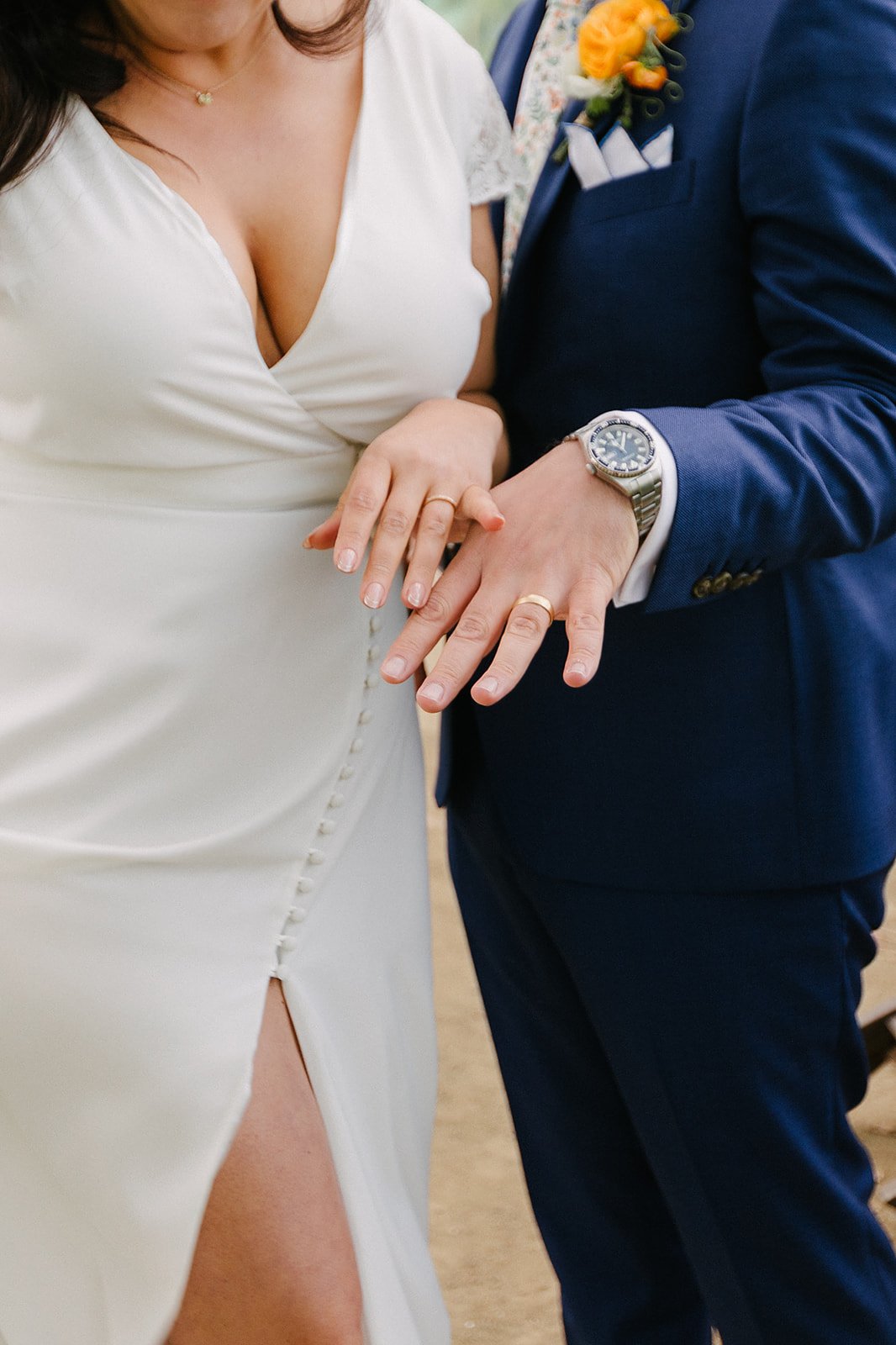Close-up of a bride and groom showing their hands with wedding rings, the bride wearing a white dress with a buttoned side slit and the groom in a dark blue suit with a watch and a yellow boutonniere.