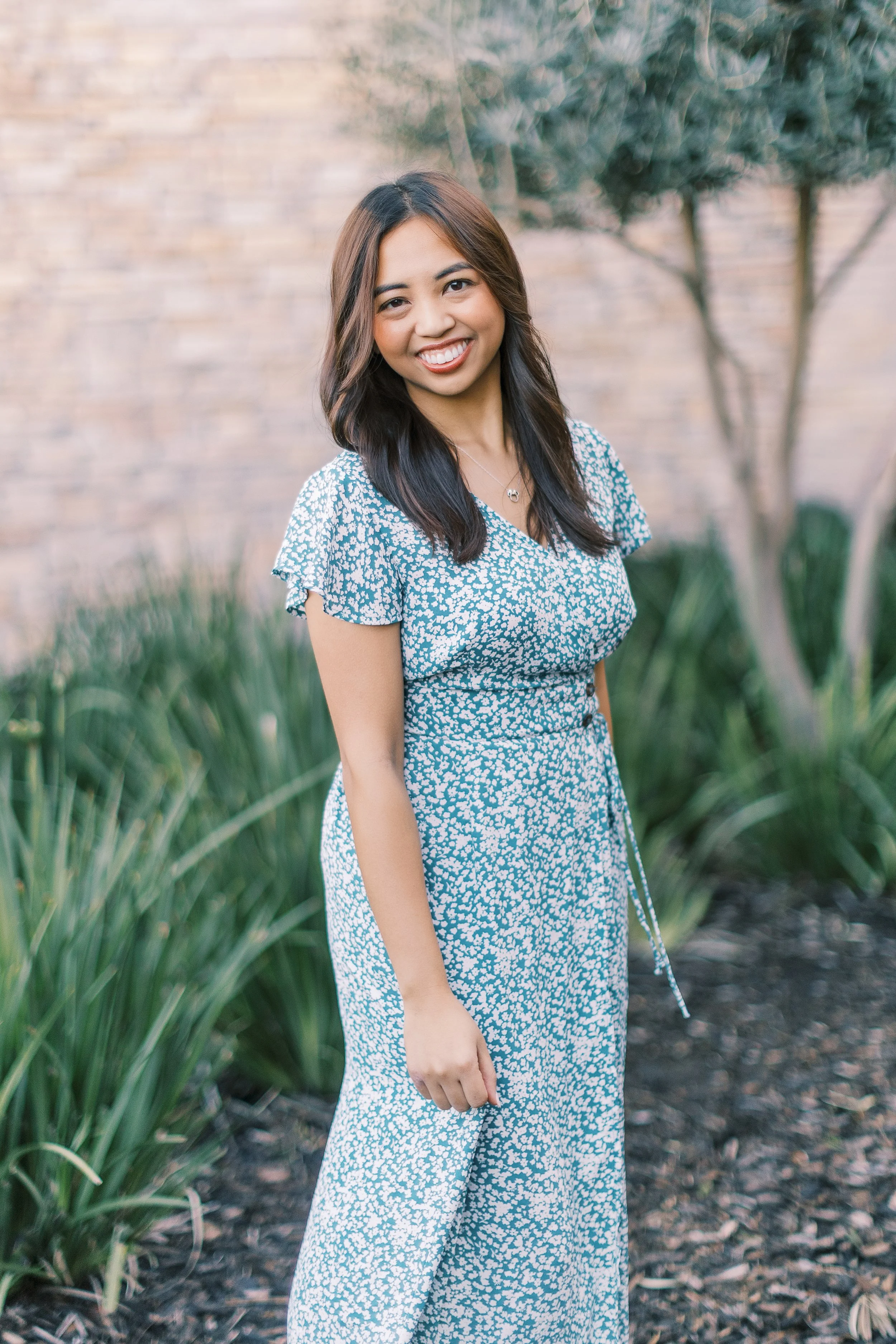 A smiling woman with long dark hair wearing a blue and white patterned dress standing outdoors near green plants and a brick wall.