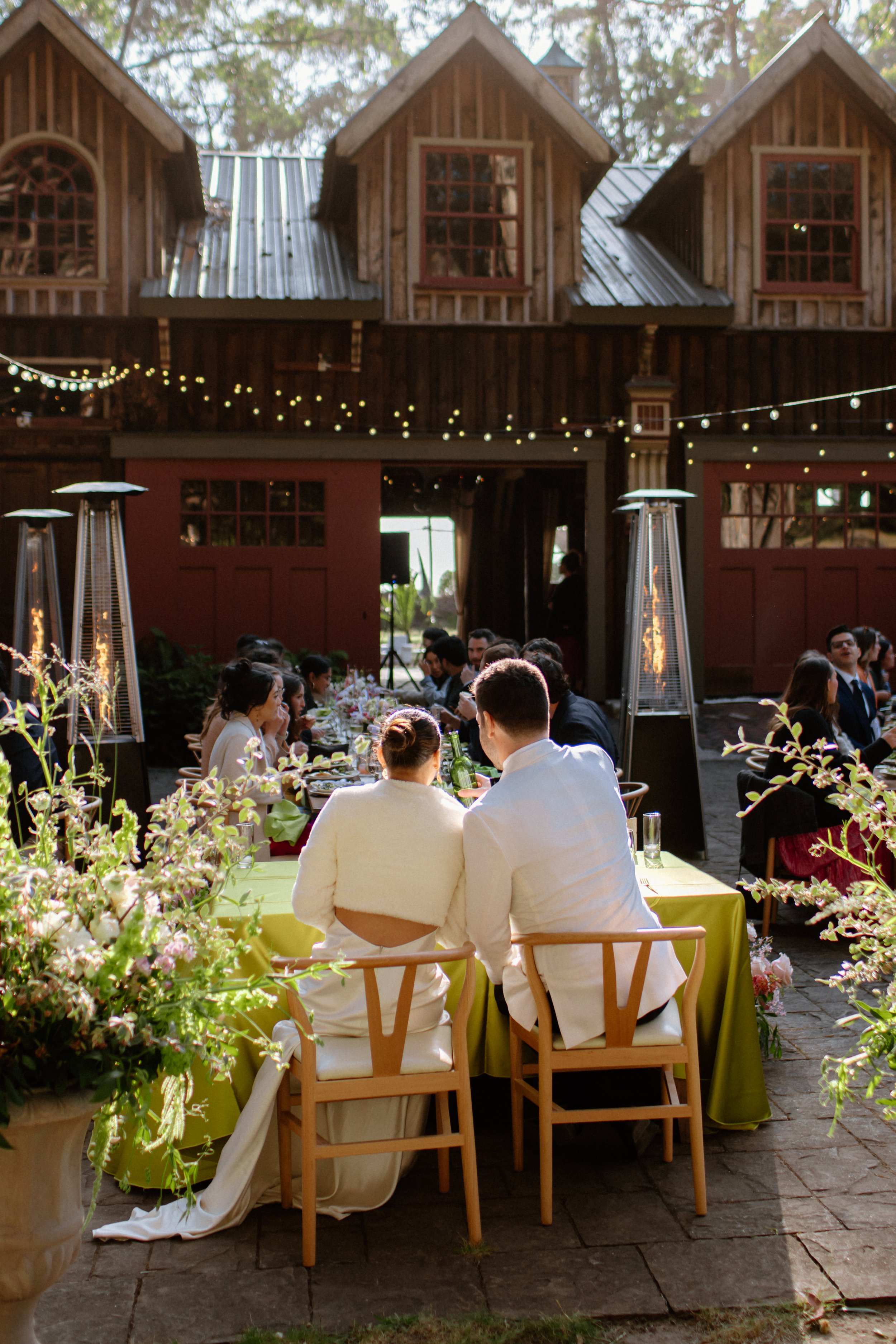 Guests at an outdoor wedding reception dining at decorated tables with string lights above and a rustic barn in the background.