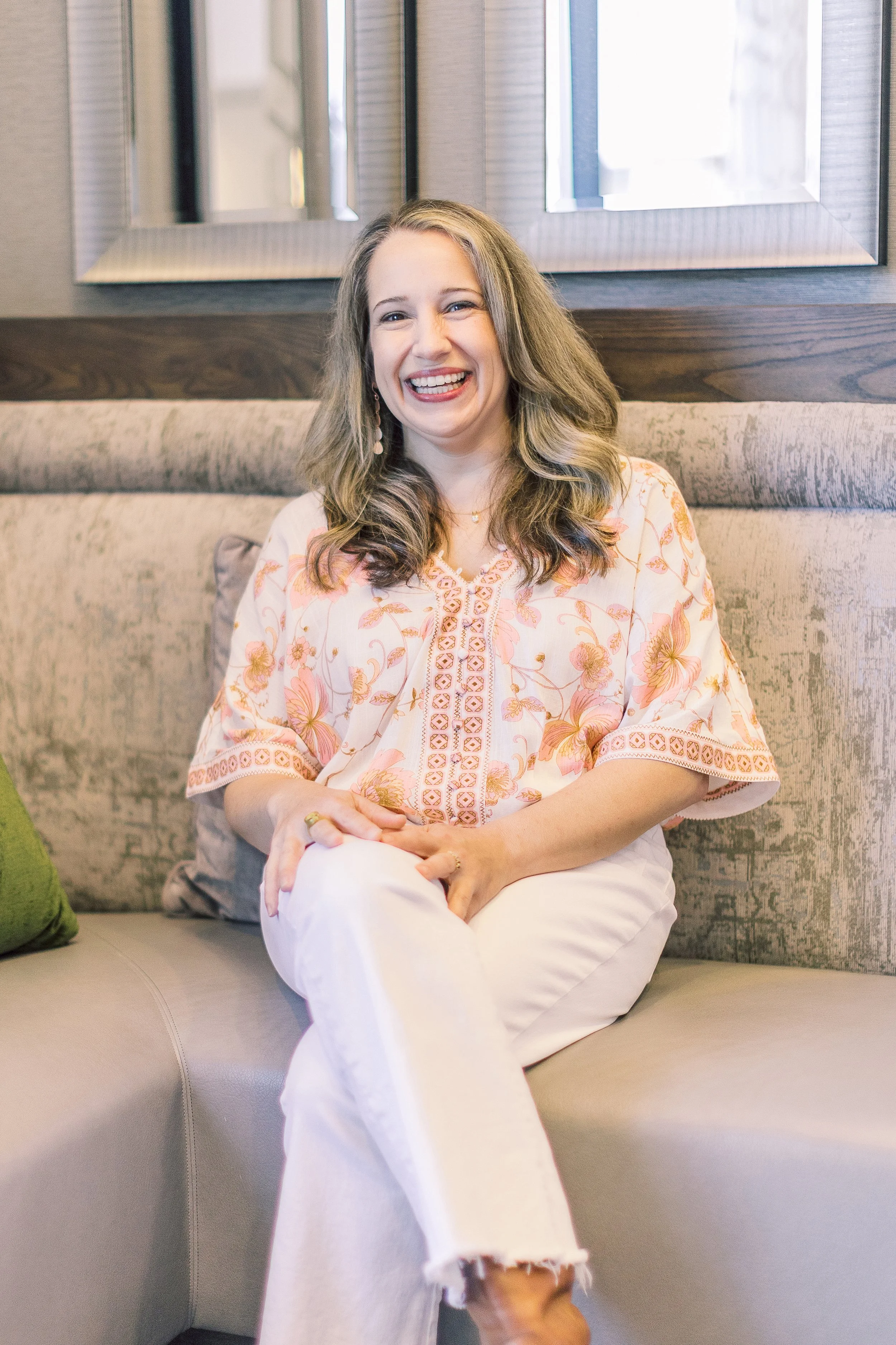 A woman with long, wavy brown hair sitting on a beige couch, smiling, wearing a cream-colored blouse with pink floral embroidery.