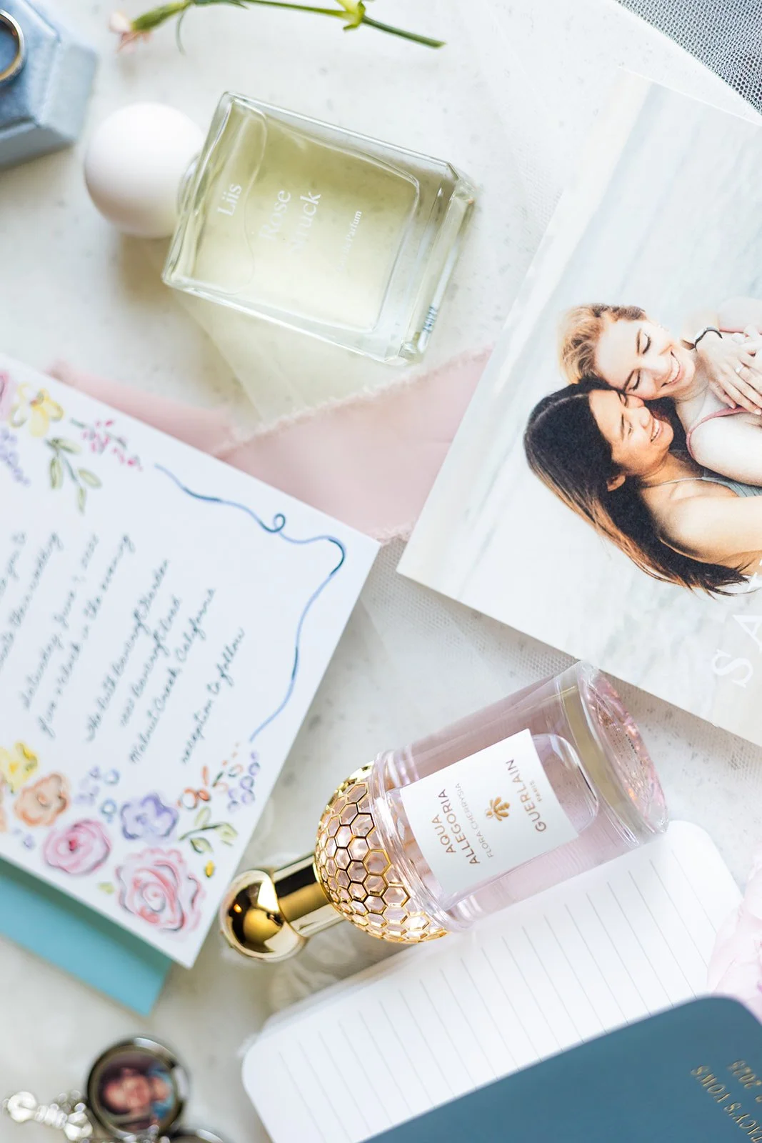 A flat lay photo of personal items, including two bottles of perfume, a photograph of two women smiling and hugging, a colorful handwritten letter with floral decorations, a small motivational card, a notebook, and a round photo locket.