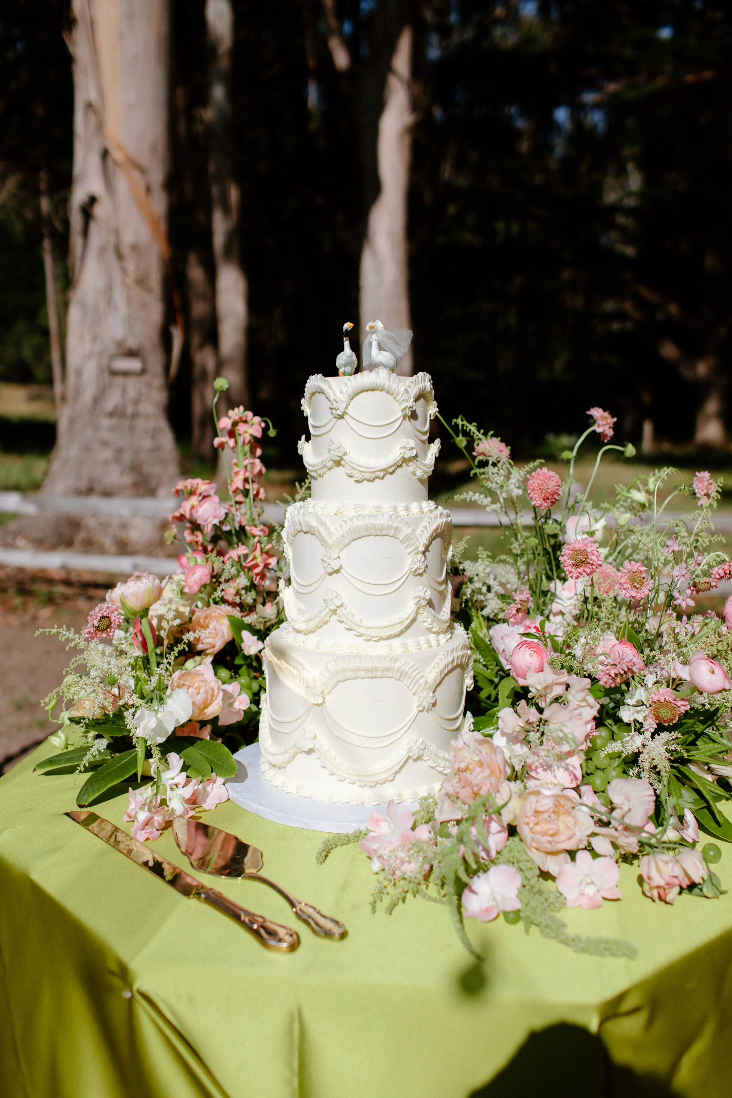 A four-tier white wedding cake with decorative piping and swags, topped with two swans, is placed on a table covered with a green tablecloth. The table is surrounded by a large bouquet of pink and white flowers, and a cake knife and server are laid beside it. The background features large trees and a natural outdoor setting.