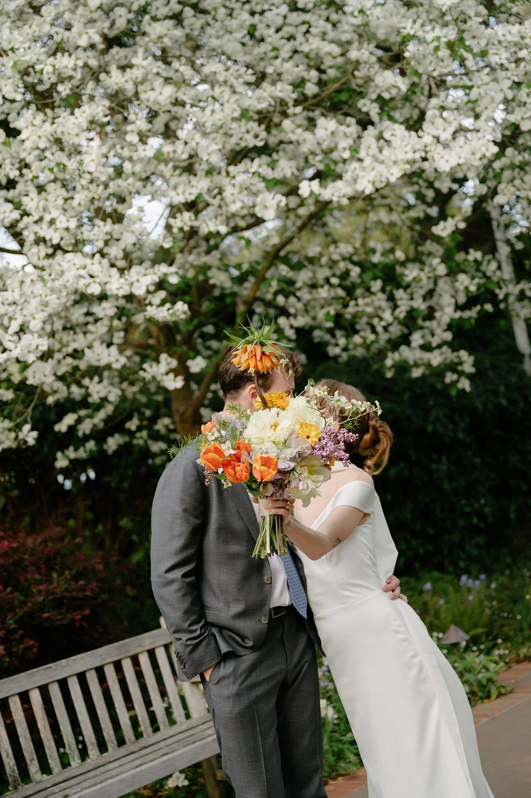 Bride and groom sharing a kiss under blooming trees, with the bride holding a large colorful bouquet.