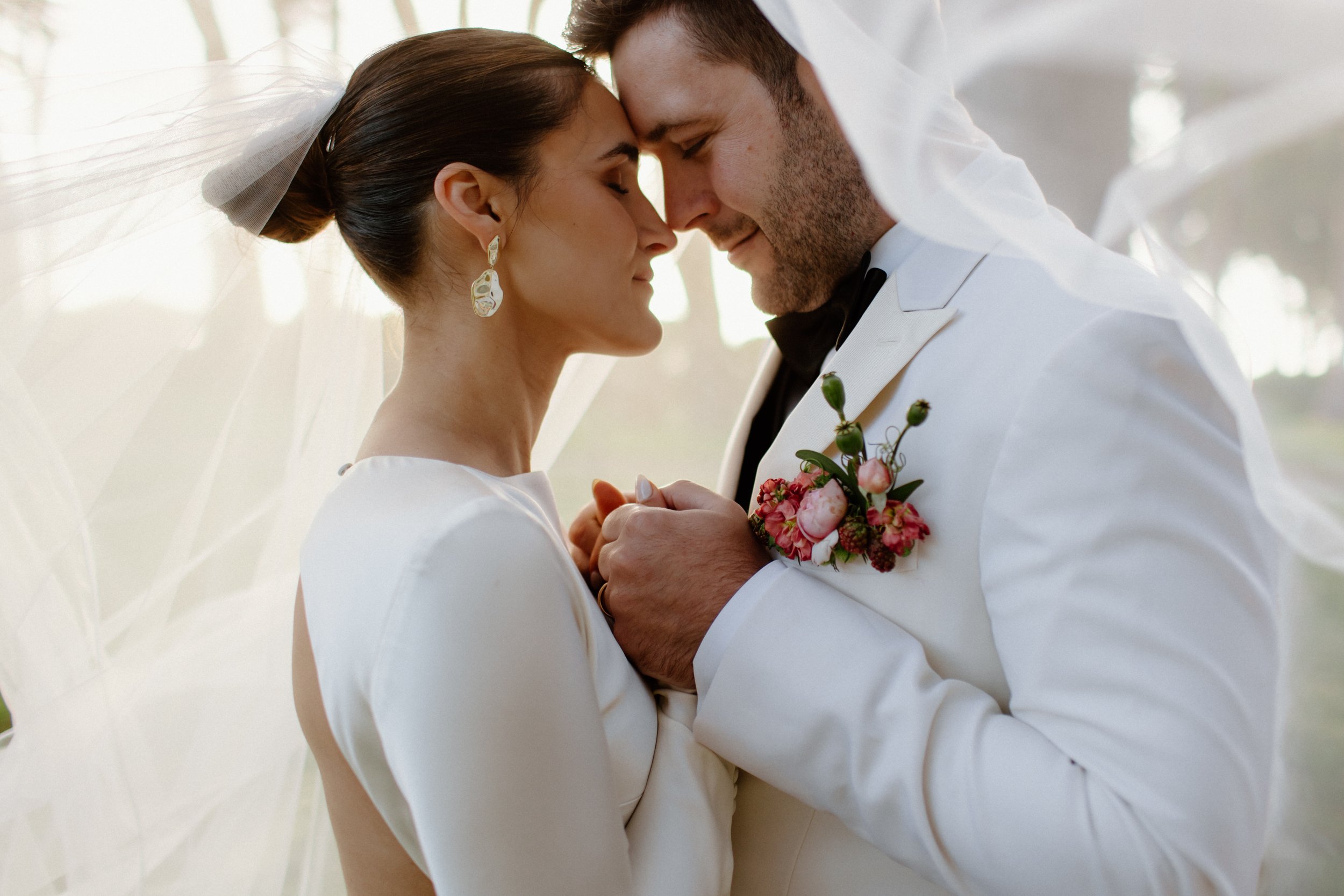 A bride and groom sharing a close moment on their wedding day, with their foreheads touching and eyes closed, surrounded by soft light.