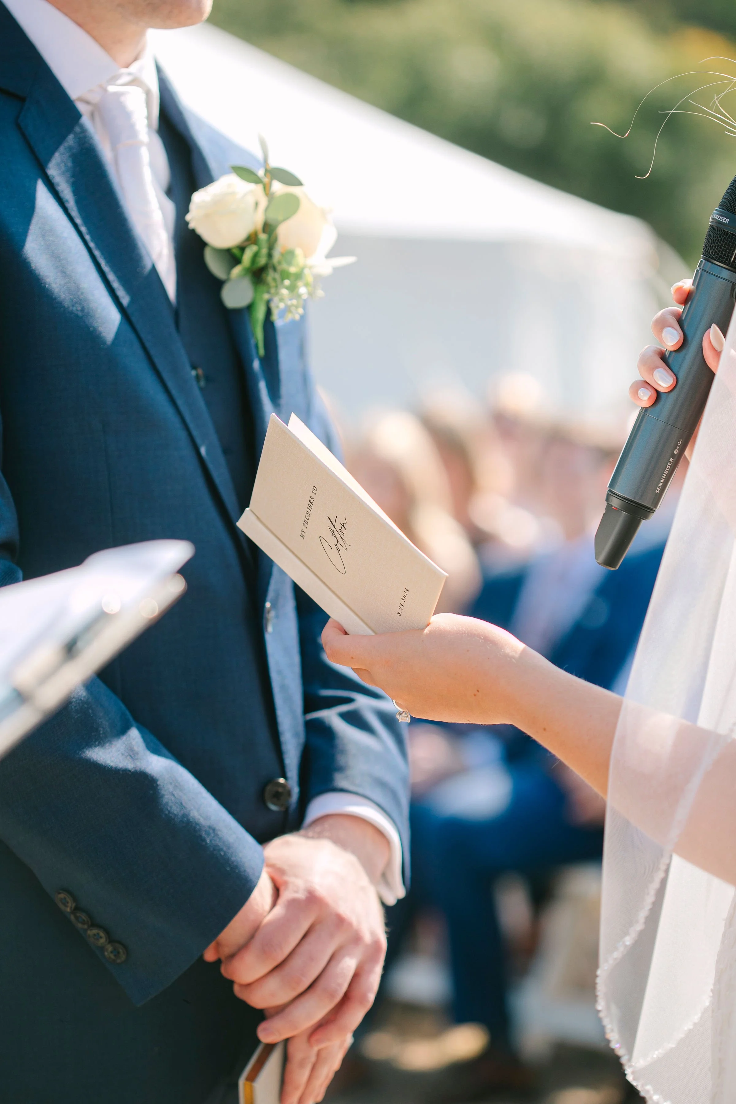 Close-up of a wedding ceremony, showing a groom in a blue suit with a white boutonniere, standing with his hands clasped as a woman in a wedding dress reads vows from a booklet and holds a microphone.