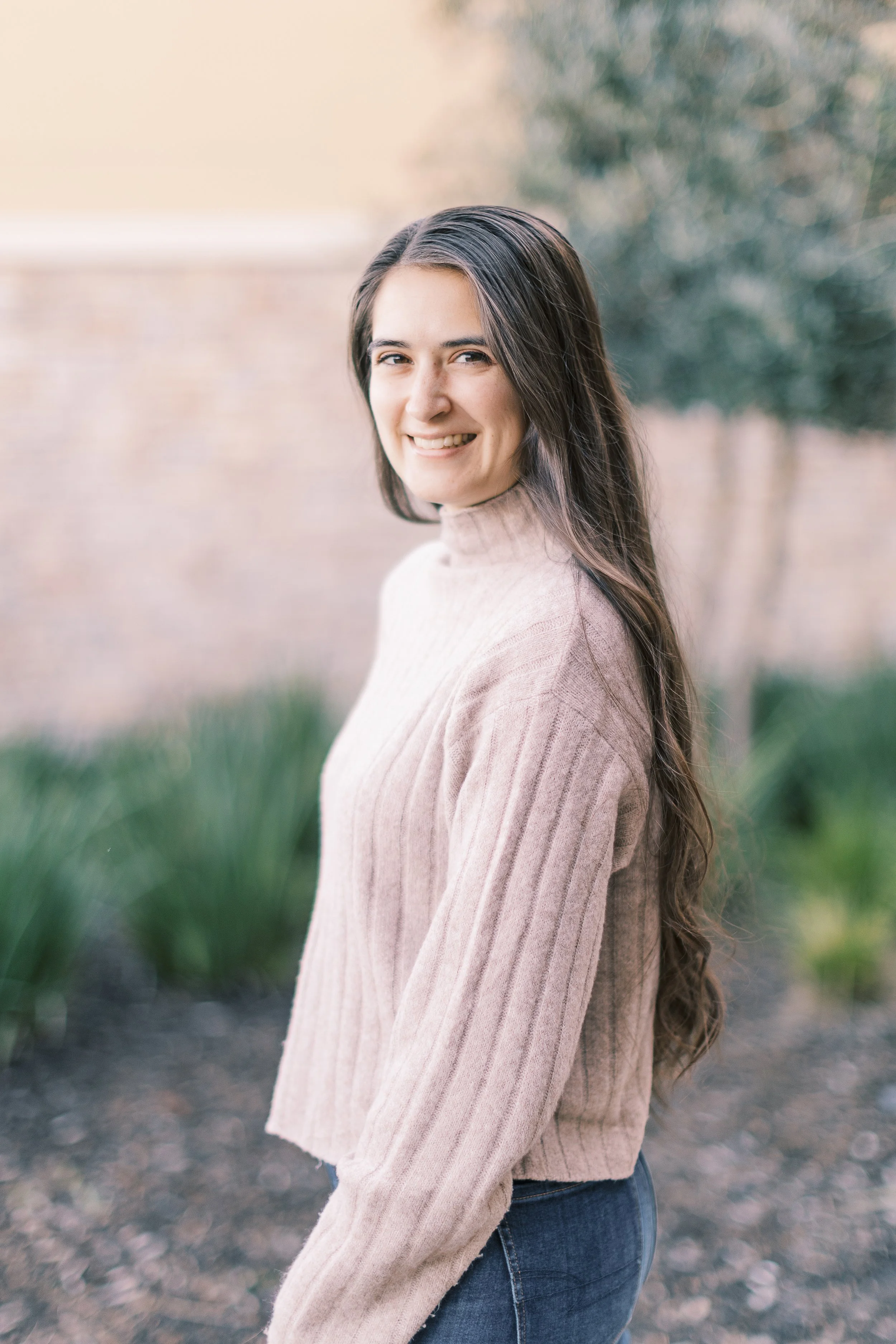 A young woman with long dark hair wearing a beige turtleneck sweater and dark jeans, standing outdoors with a blurred background of greenery and a brick wall, smiling at the camera.