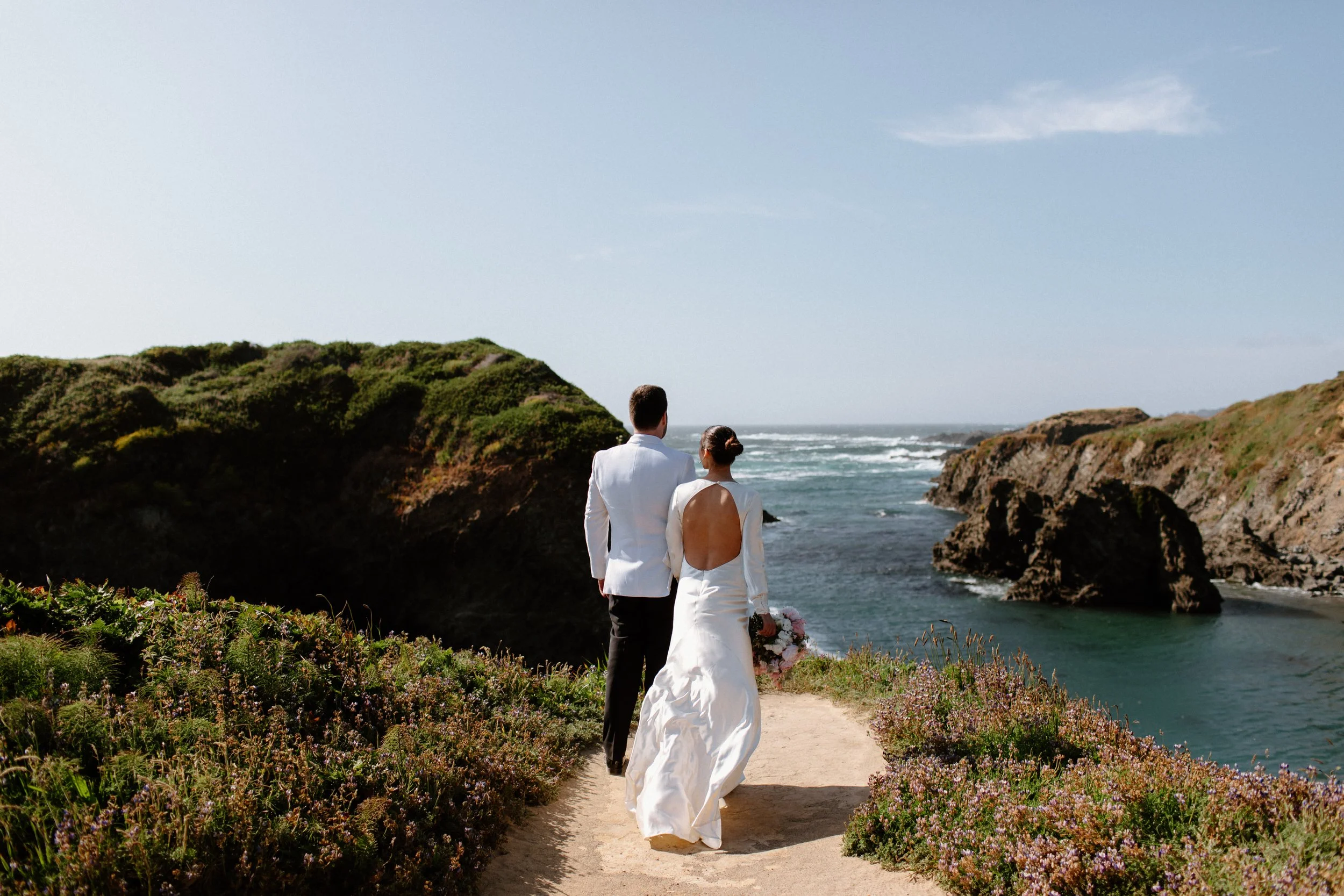 Bride and groom walk along a coastal path with rocky cliffs and ocean waves in the background on a clear day