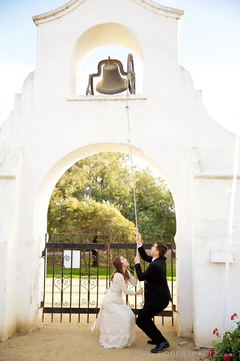A couple dressed in wedding attire, a woman in a white lace wedding dress and a man in a black tuxedo, are ringing a large outdoor church bell.